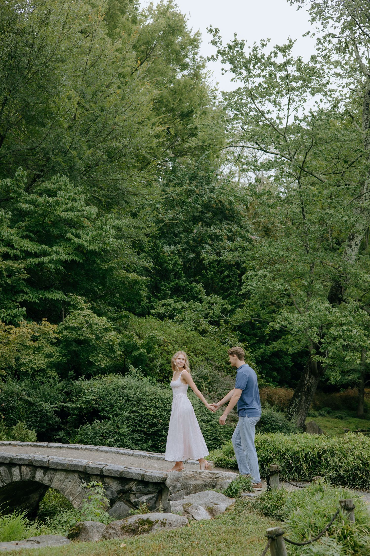 A couple holding hands while crossing a stone bridge in a lush garden setting.