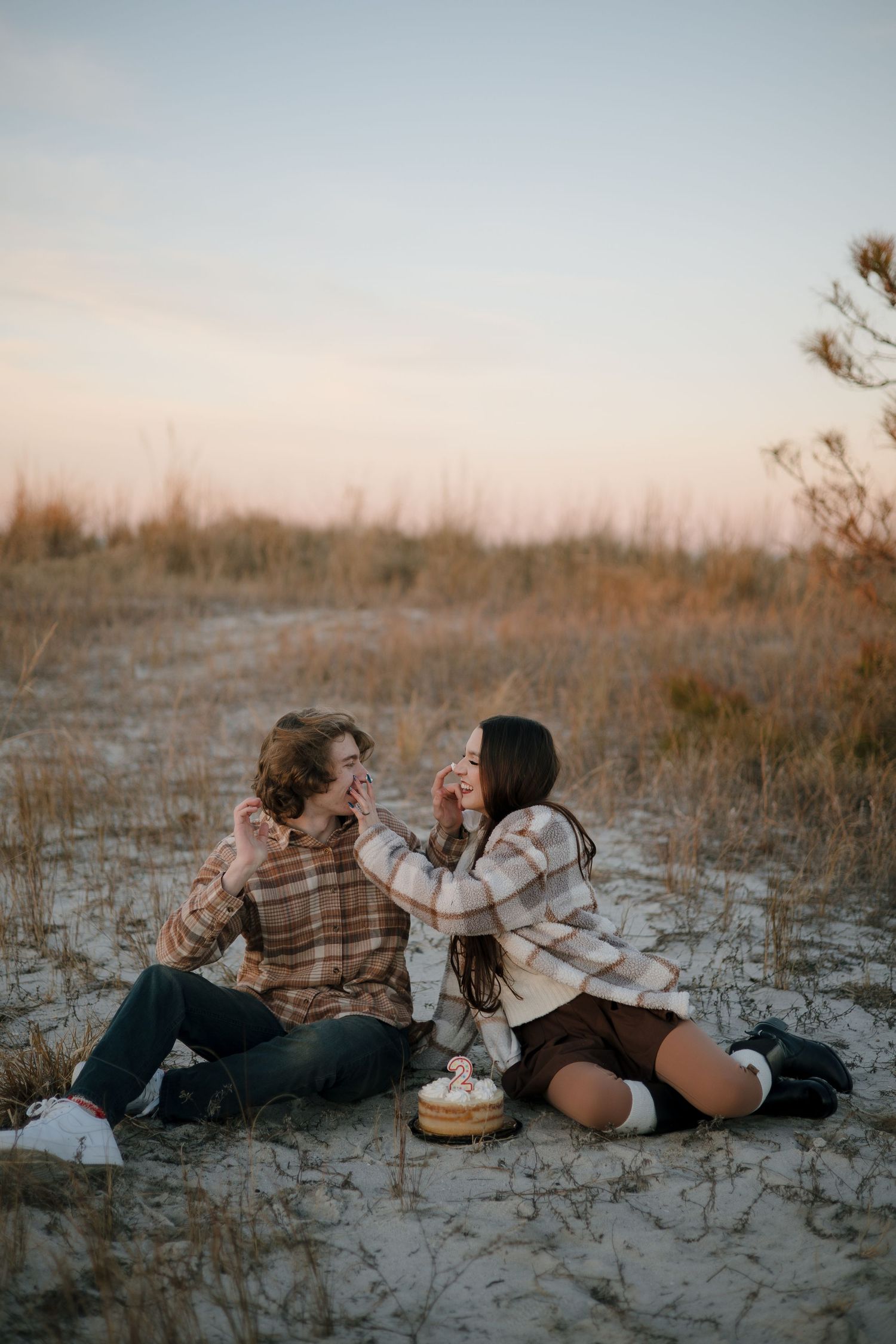A couple shares a cozy winter picnic on a beach at sunset, wrapped in plaid blankets with a pie between them.