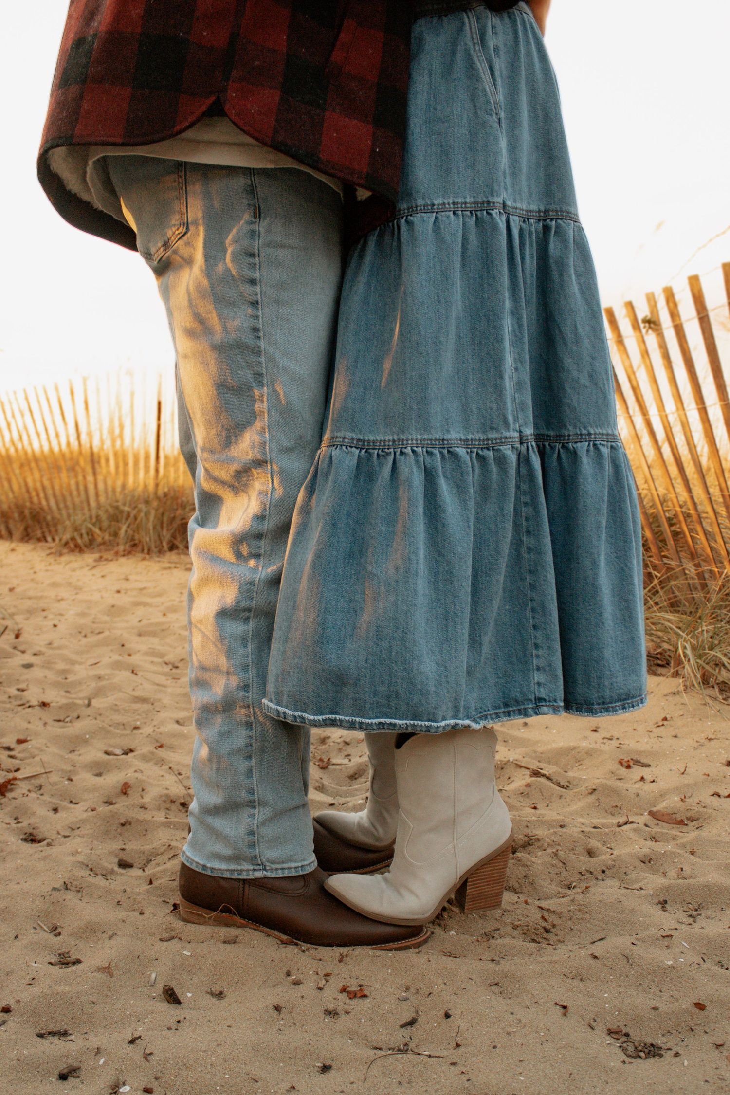 Close-up of denim jeans and cowboy boots standing on sandy beach near wooden fence at sunset.
