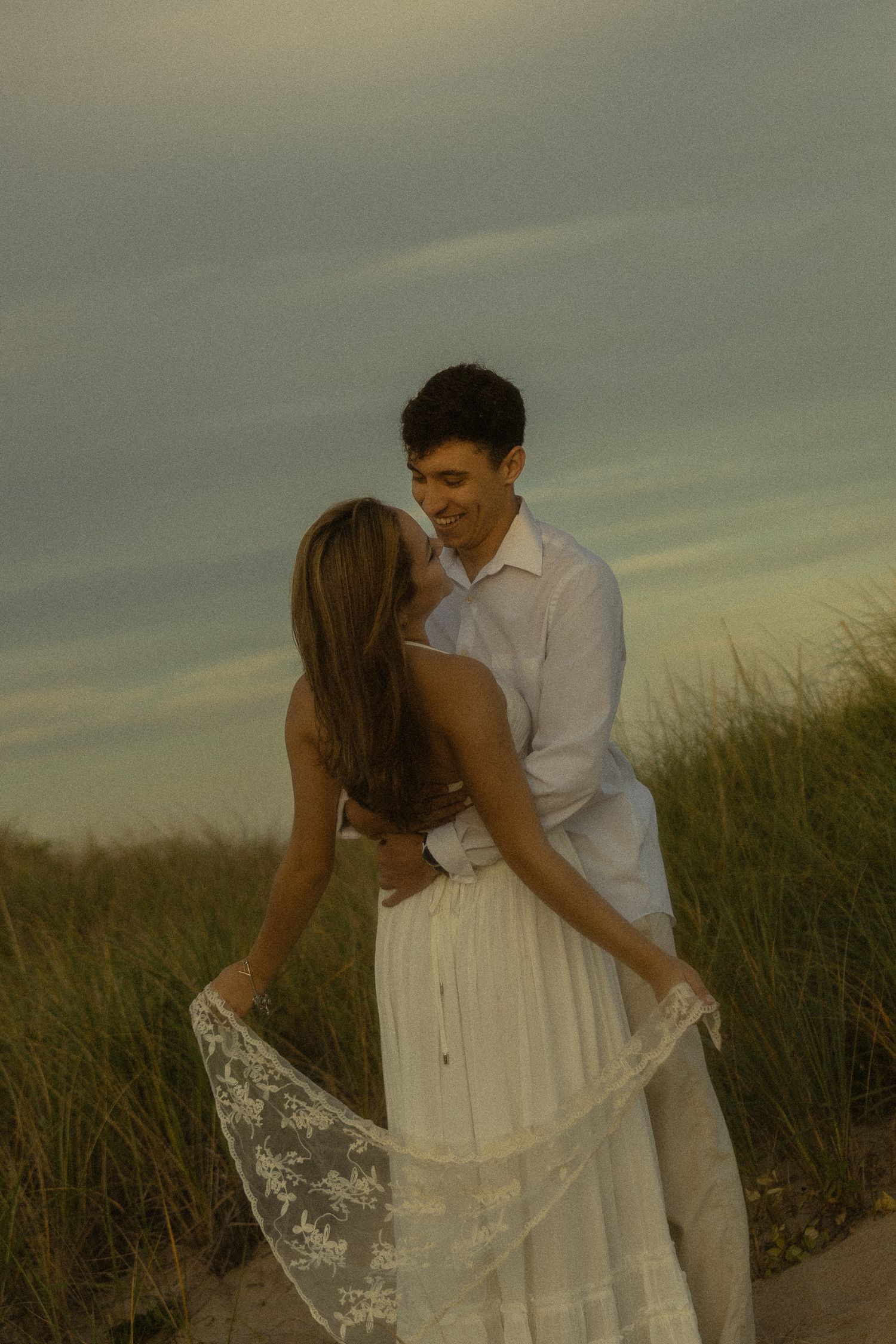 Couple in white outfits embracing on a beach path with tall grass and cloudy sky at golden hour.