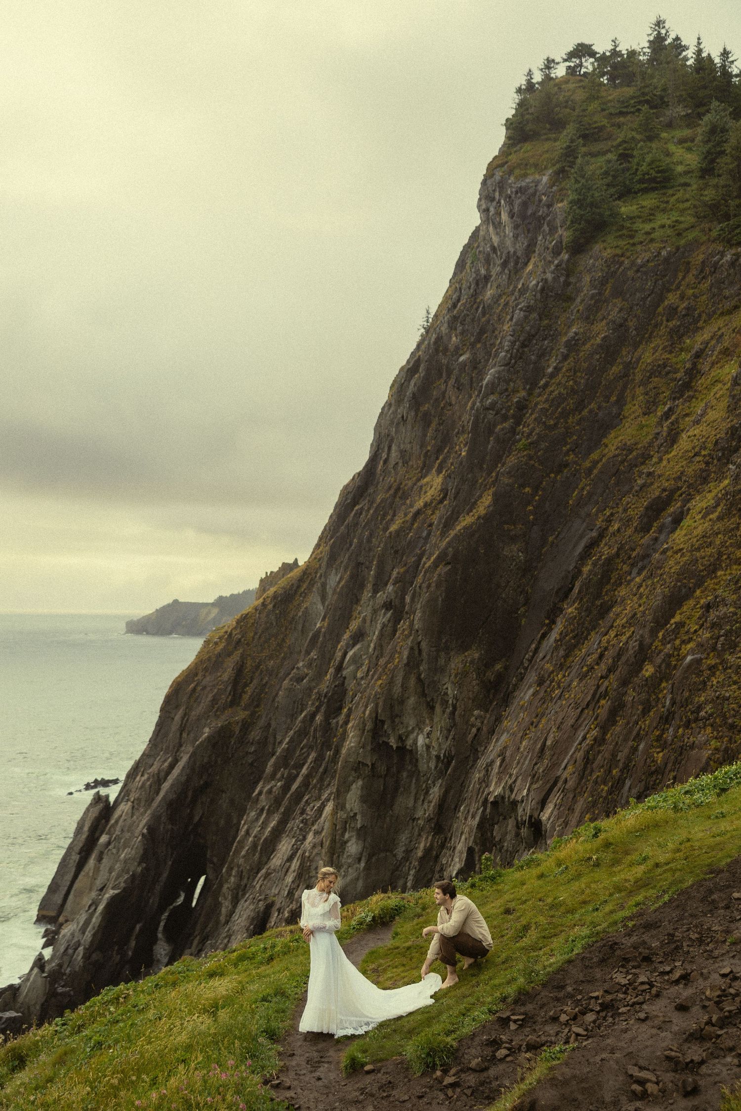 Bride and groom pose on a dramatic coastal cliff overlooking the ocean on a cloudy day.