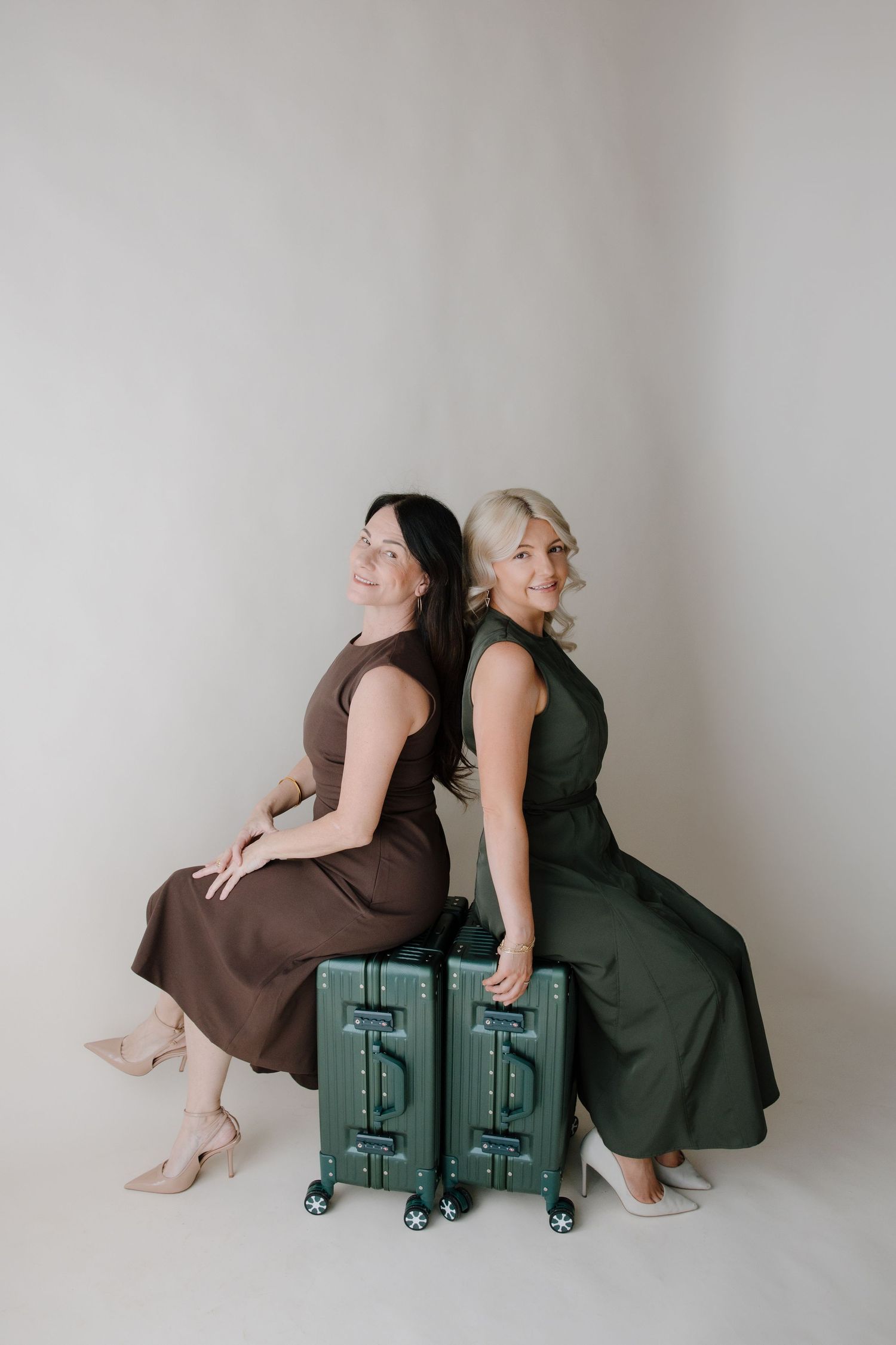 Two people in contrasting brown and black dresses pose playfully on matching green suitcases against a neutral backdrop.