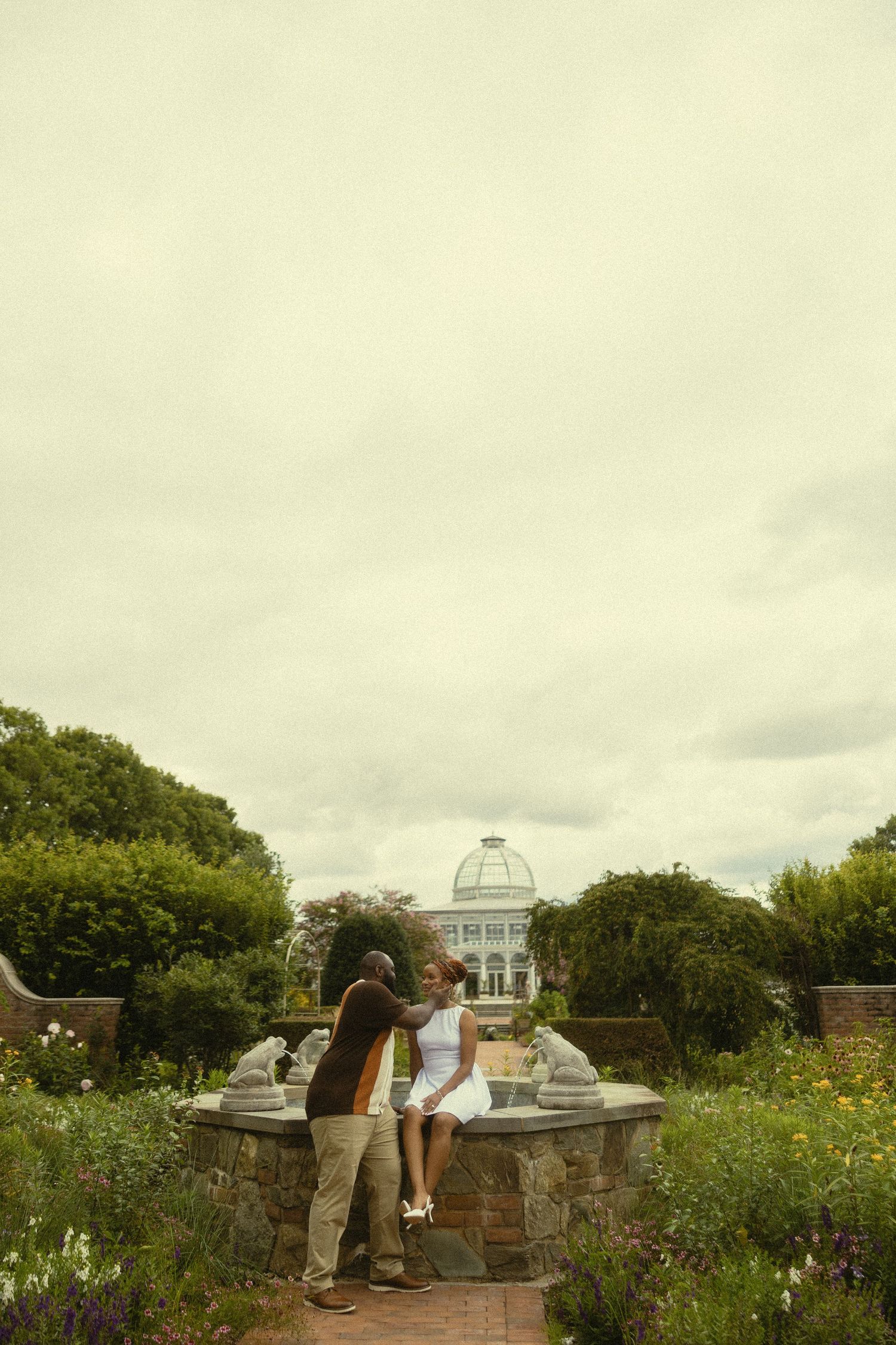 Couple sharing a romantic moment in a lush garden with a historic greenhouse building in the background.