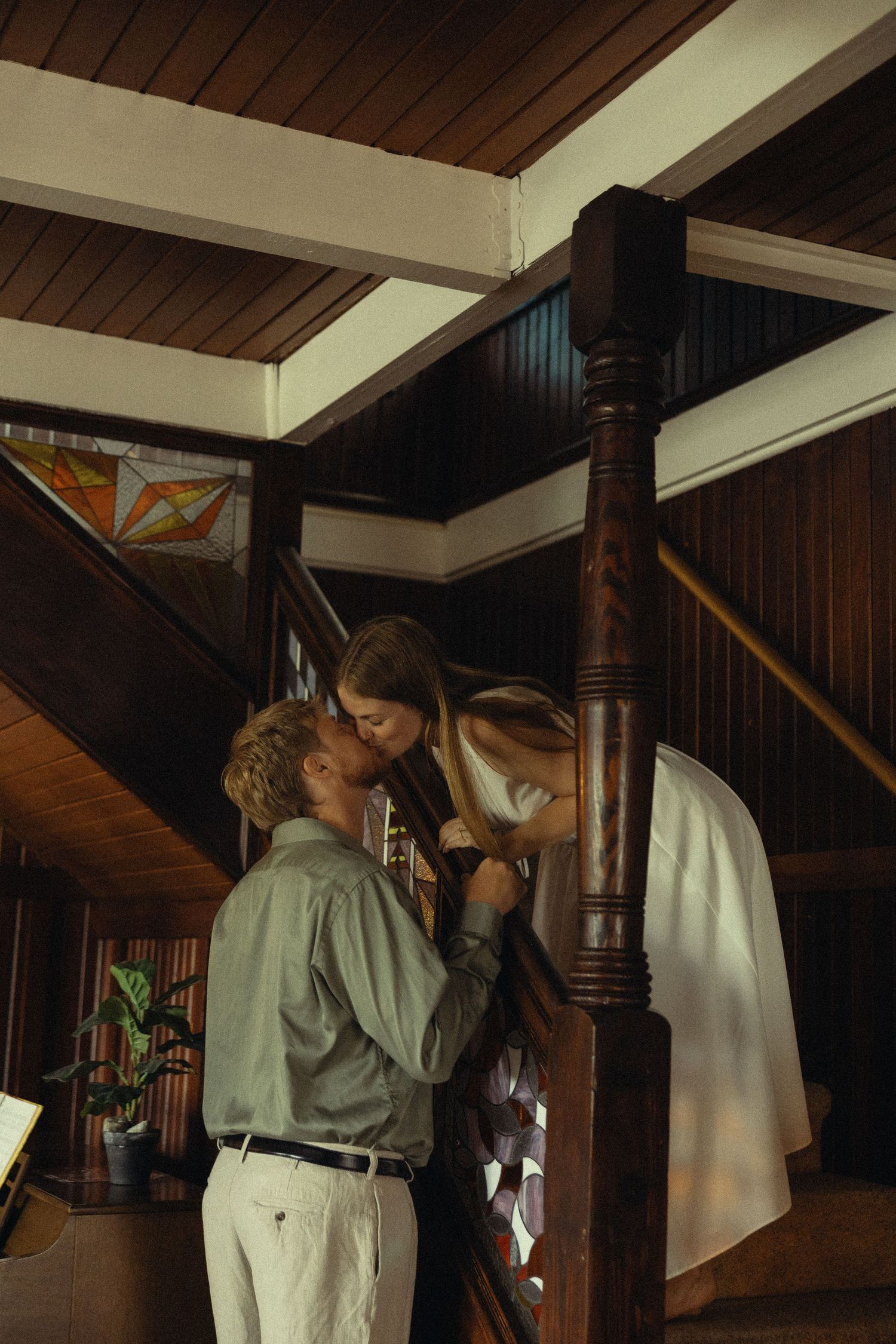 Couple sharing a romantic kiss by a wooden pillar inside a rustic interior with stained glass windows.