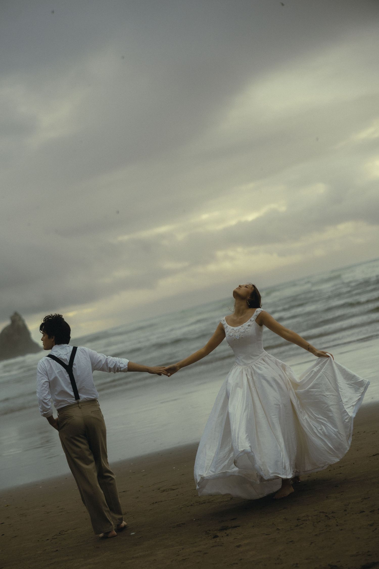 Bride in white gown and groom in suspenders hold hands on a moody beach with dramatic cloudy skies.