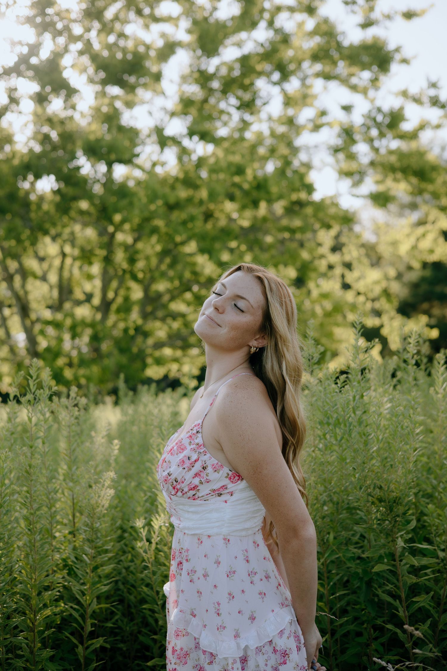 A person in a floral dress stands in a field of tall grass and trees during golden hour lighting.
