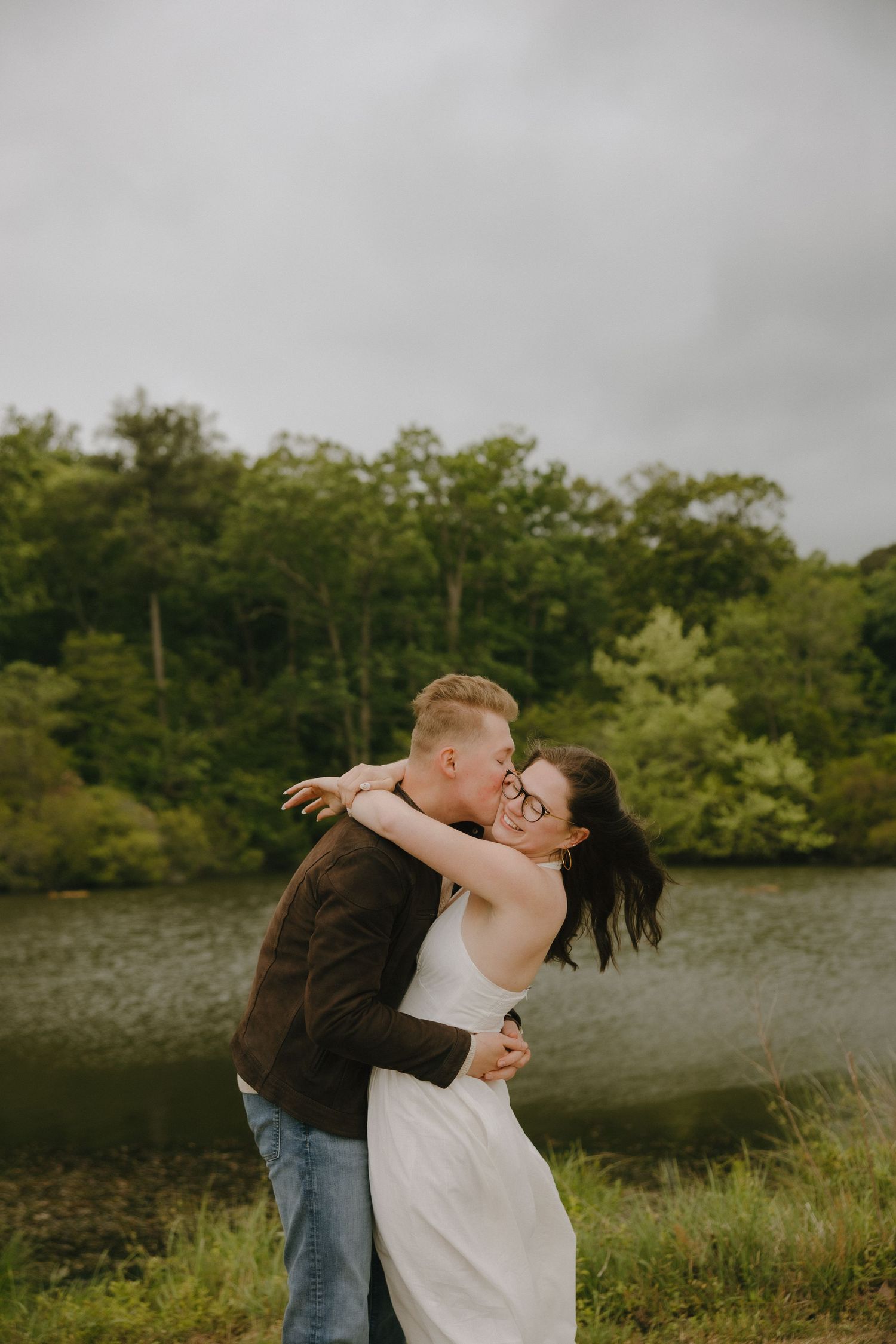 Couple embracing and kissing near a serene lake surrounded by lush green trees on a cloudy day.