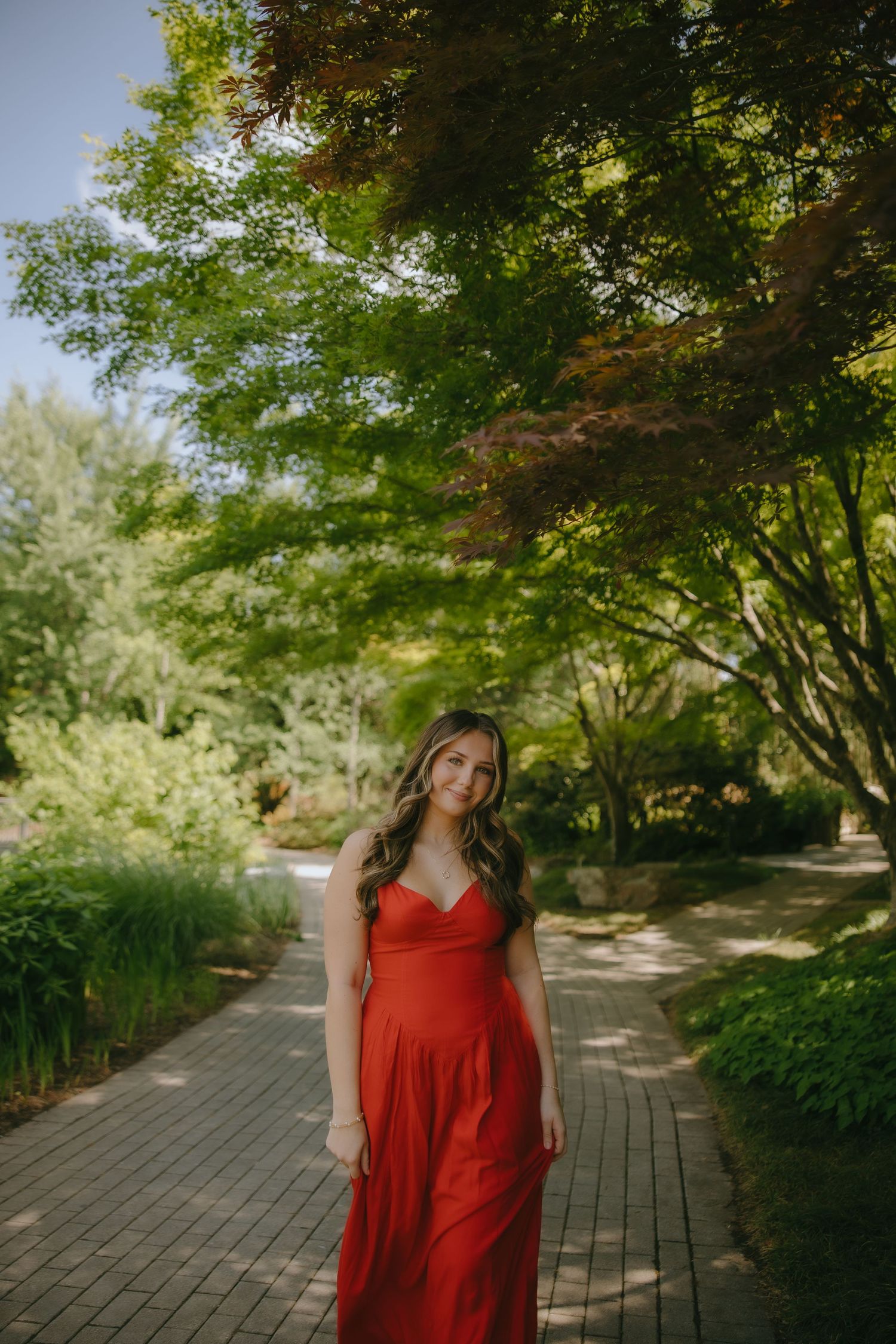 A young woman in a red maxi dress poses on a brick path surrounded by lush green trees in a garden.