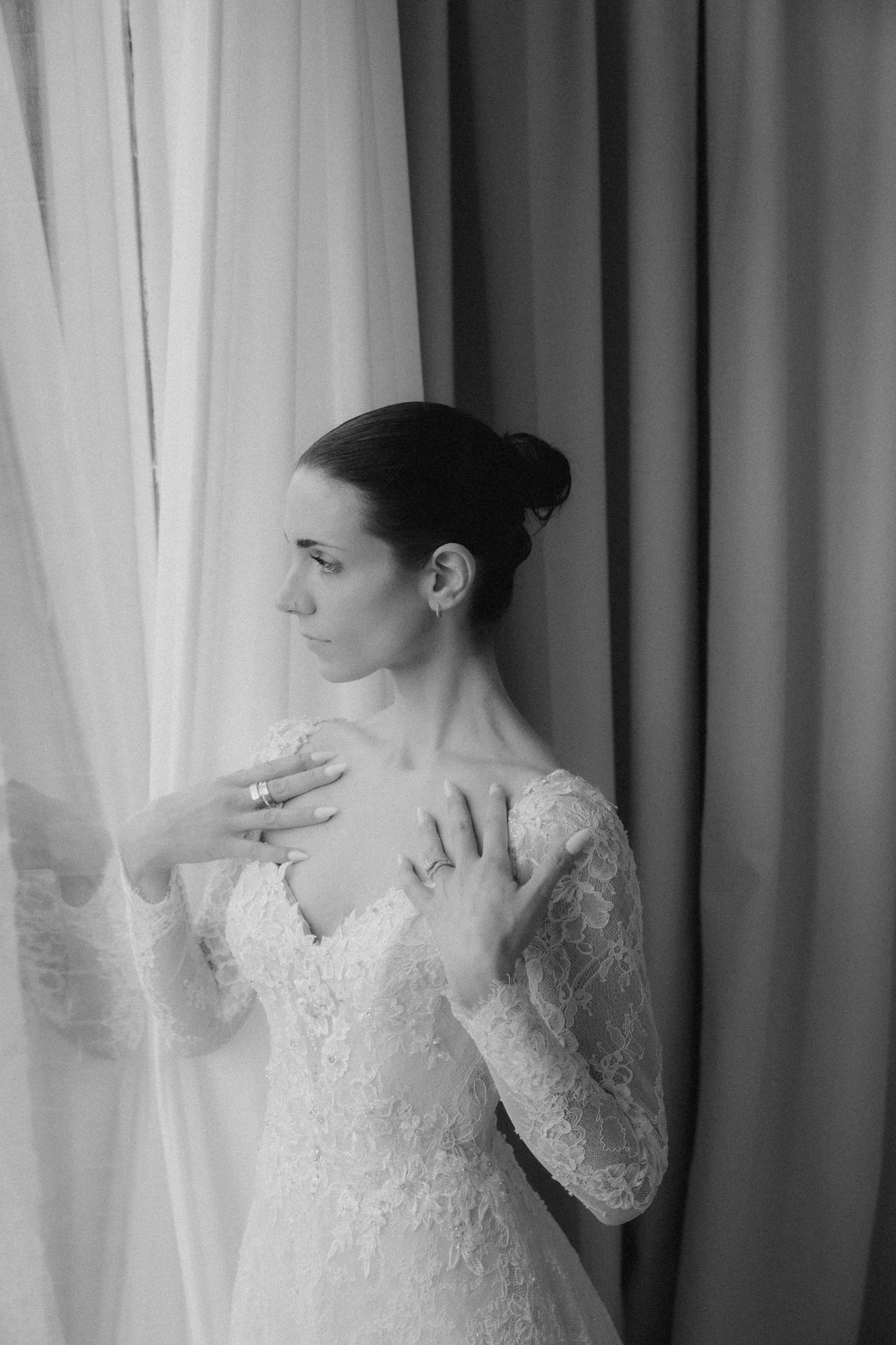 Black and white portrait of a bride in lace wedding dress standing by window, hands on chest, wearing rings.