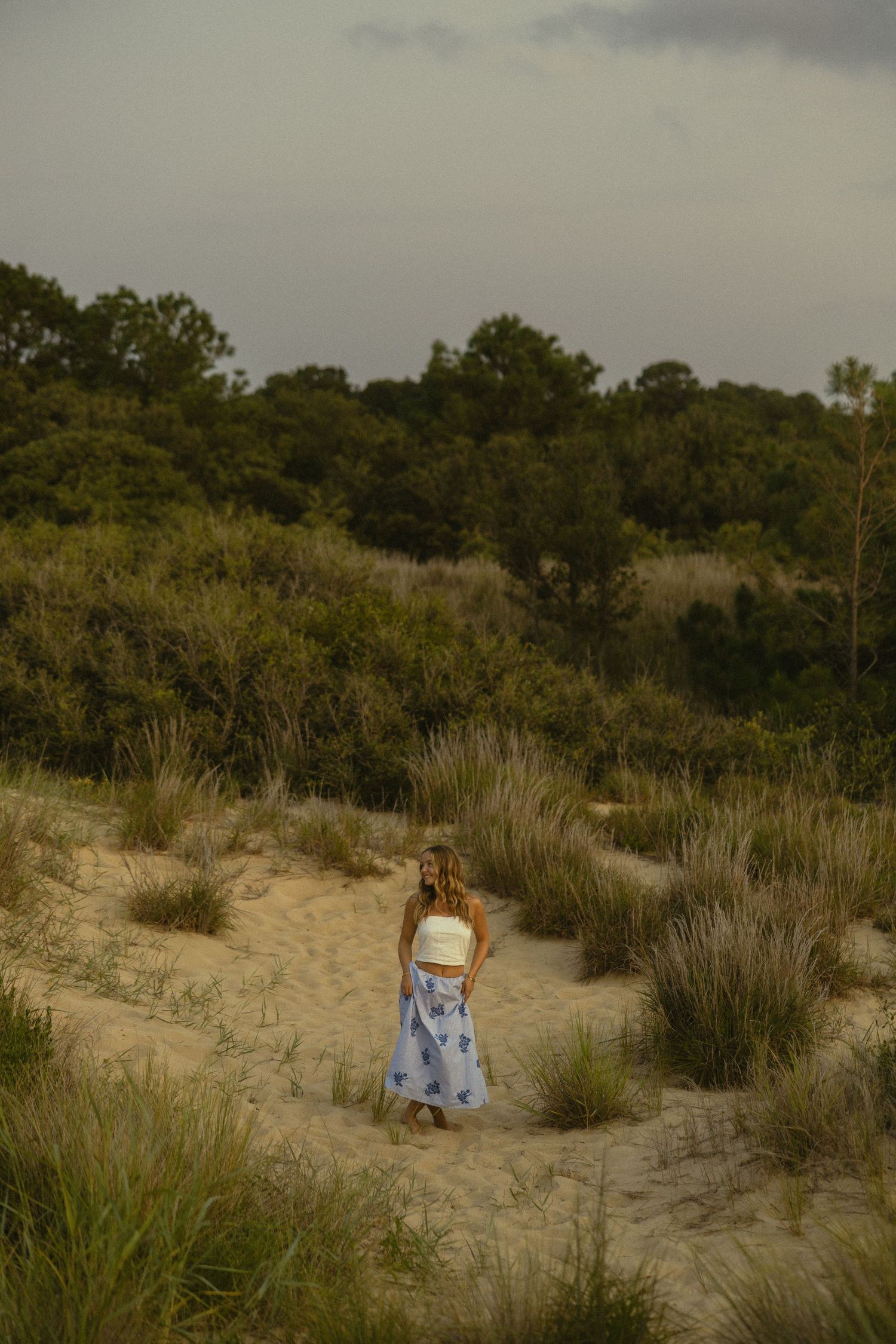 Woman in white crop top and blue floral skirt walking on sandy dunes surrounded by lush green vegetation under cloudy sky.