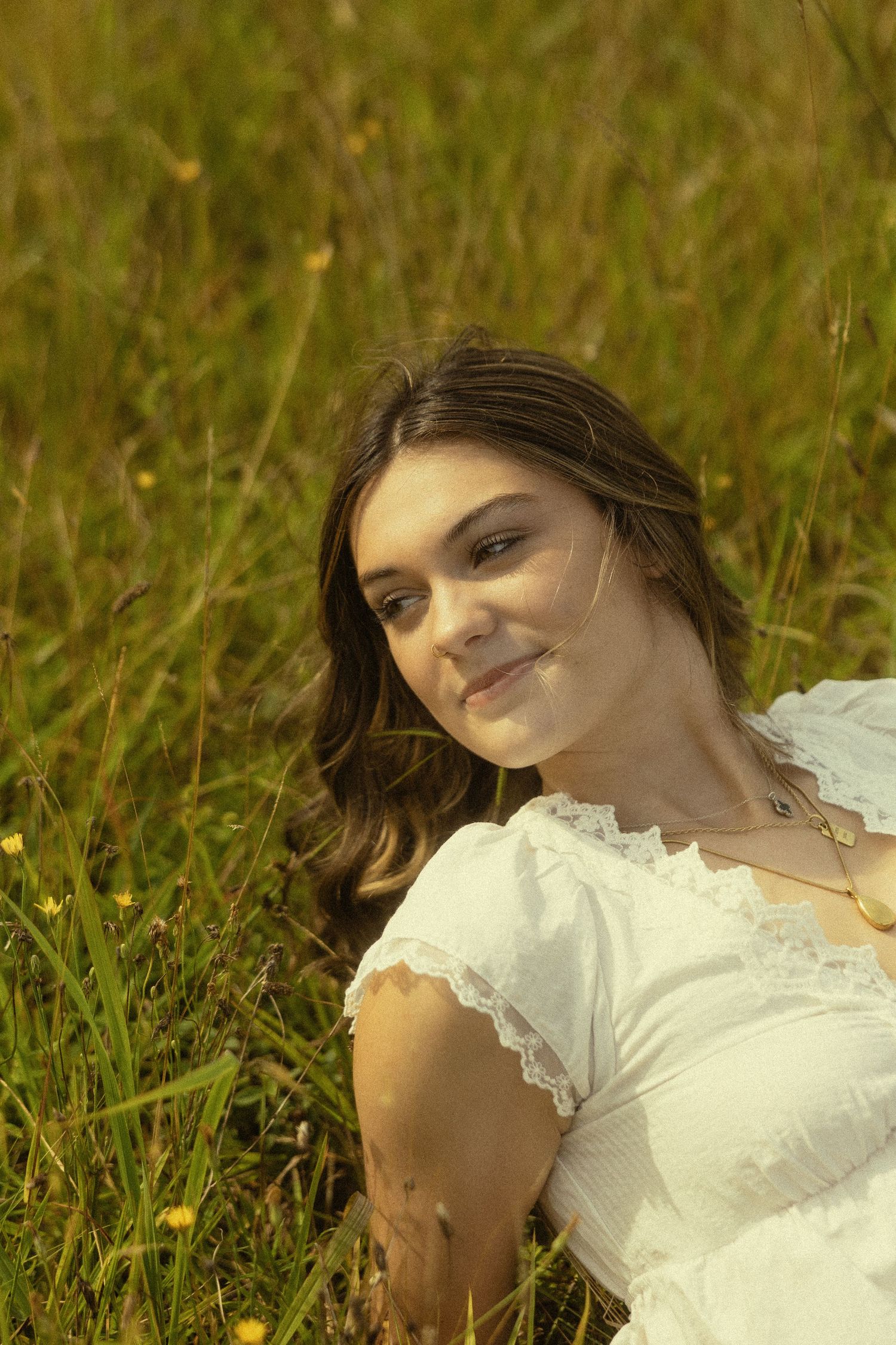 Young woman in white lace dress smiling while sitting in a sunny green meadow with wildflowers.