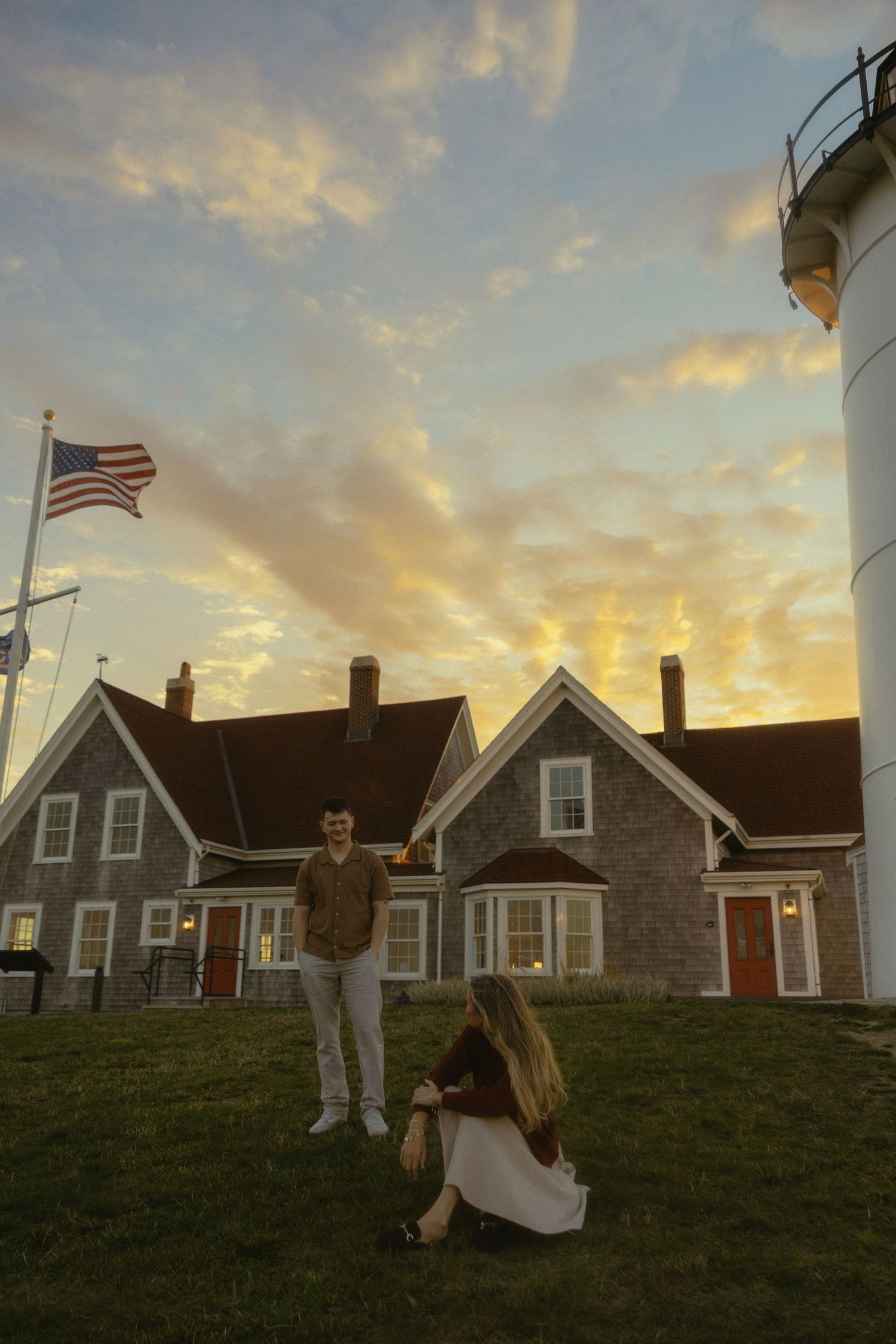 Couple at sunset near a lighthouse and colonial house with American flag flying against a dramatic golden sky.