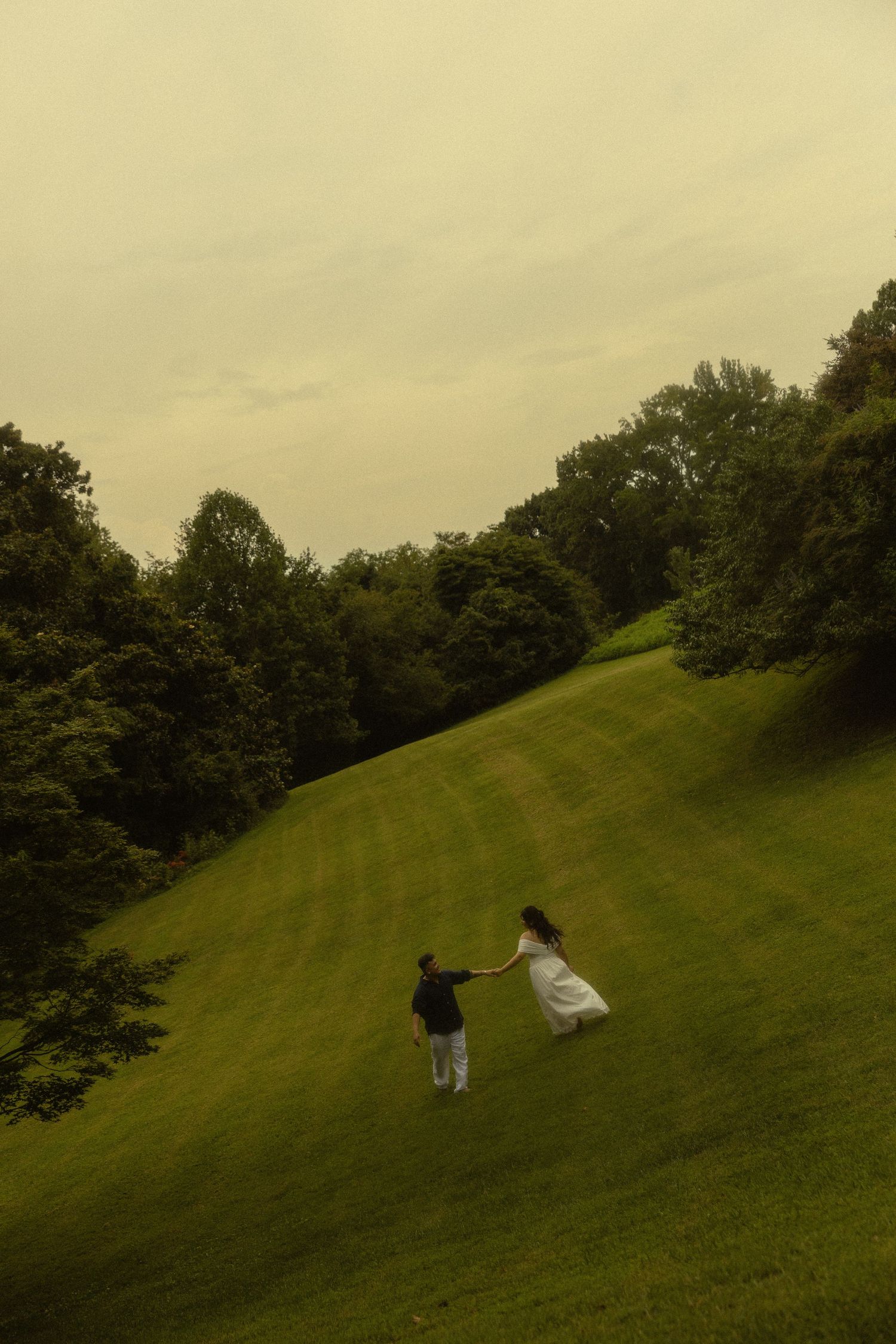 A couple holds hands while walking up a lush green hillside surrounded by trees under an overcast sky.
