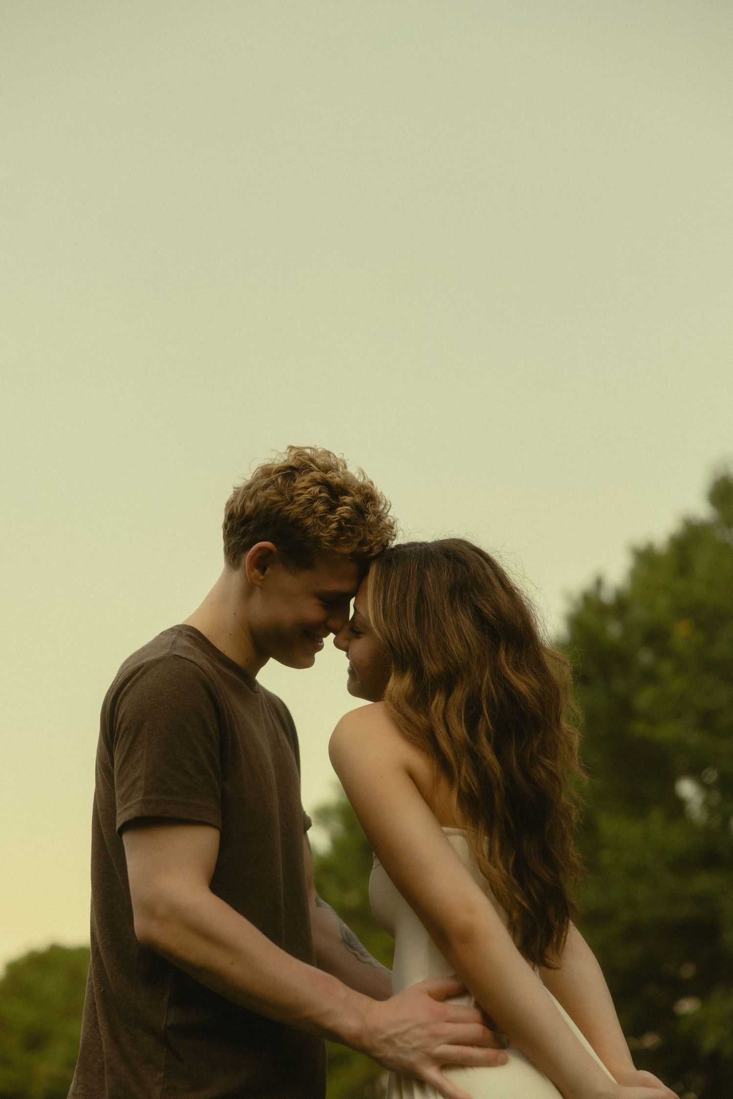 Romantic couple sharing an intimate moment outdoors, touching foreheads tenderly against a soft golden sky.