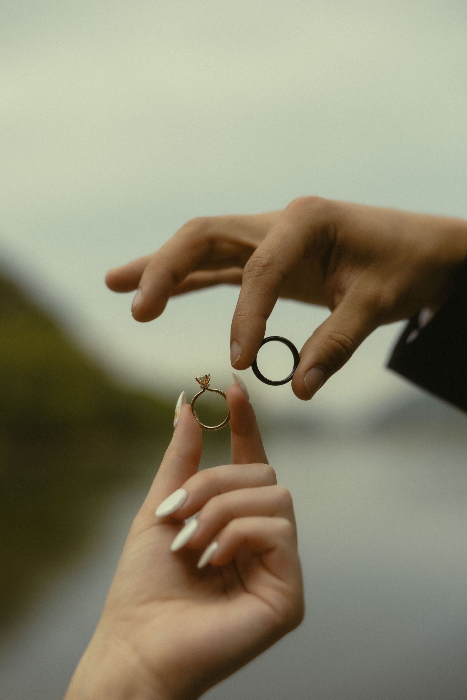 Two hands holding wedding rings outdoors, one with white nails displaying a diamond engagement ring, the other holding a plain band.
