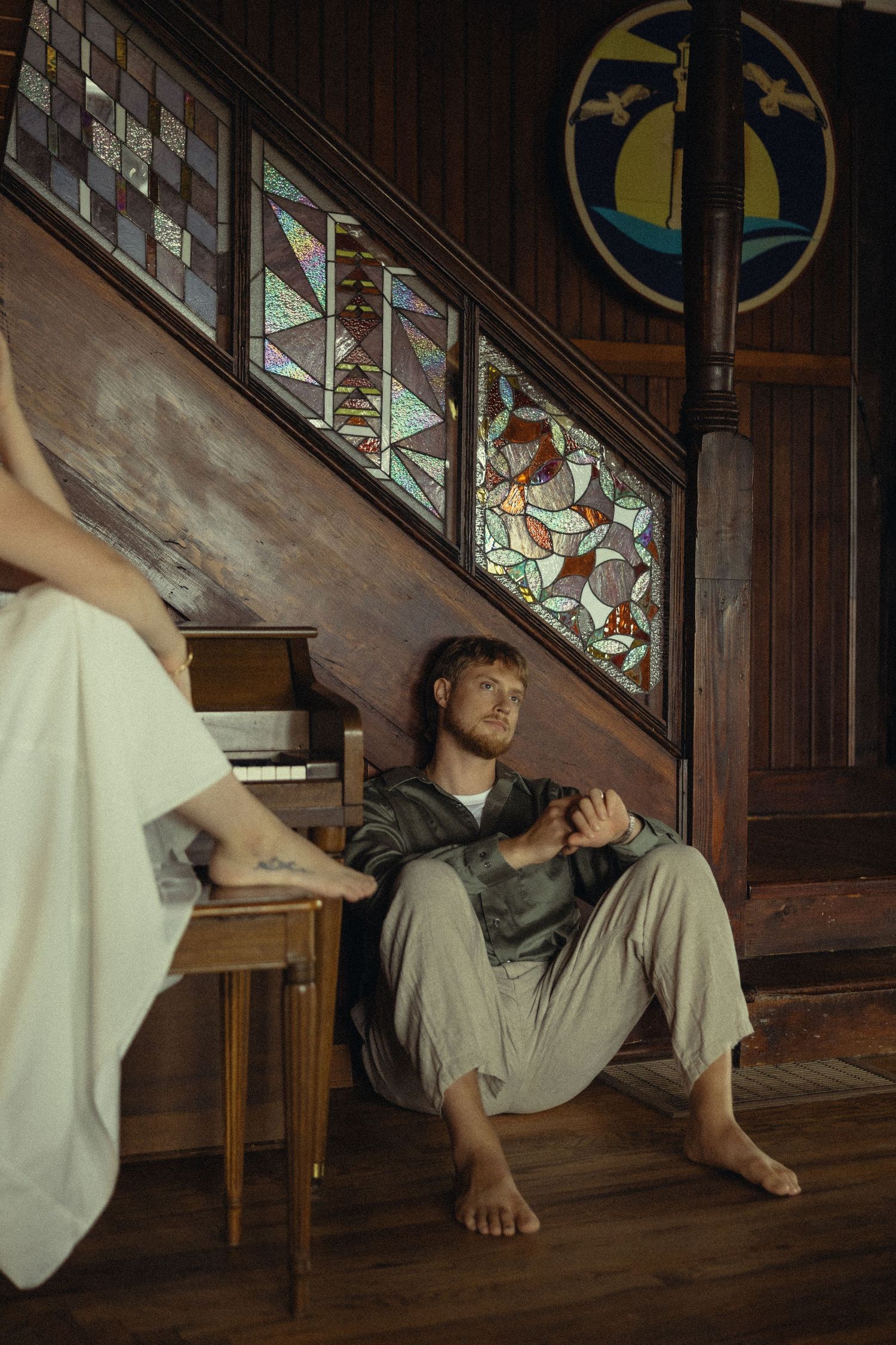 Man sitting barefoot on wooden floor by stained glass staircase, relaxing near a piano in a rustic interior.