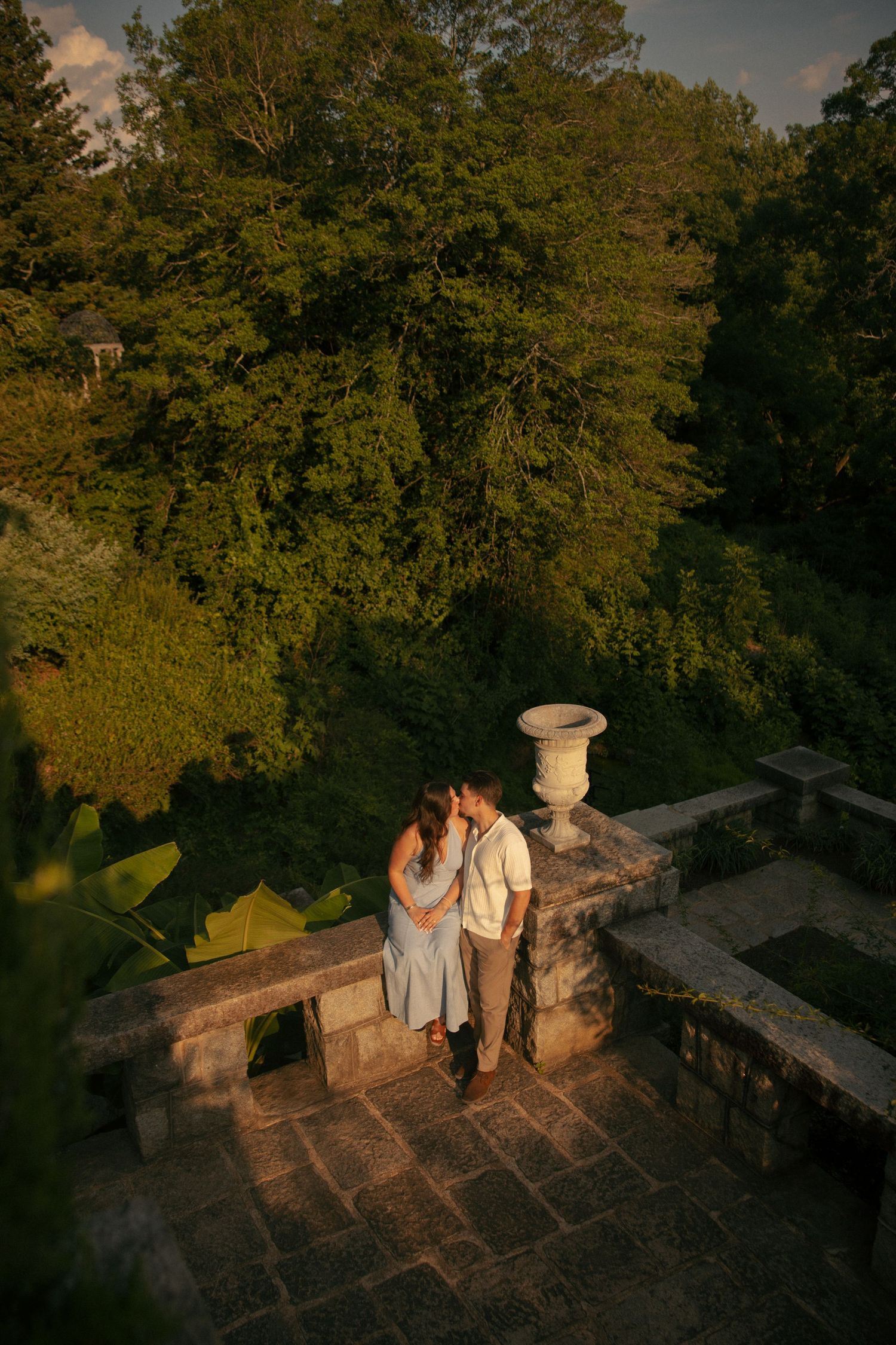 Couple embracing on a stone terrace surrounded by lush green trees during golden hour at an elegant garden venue.