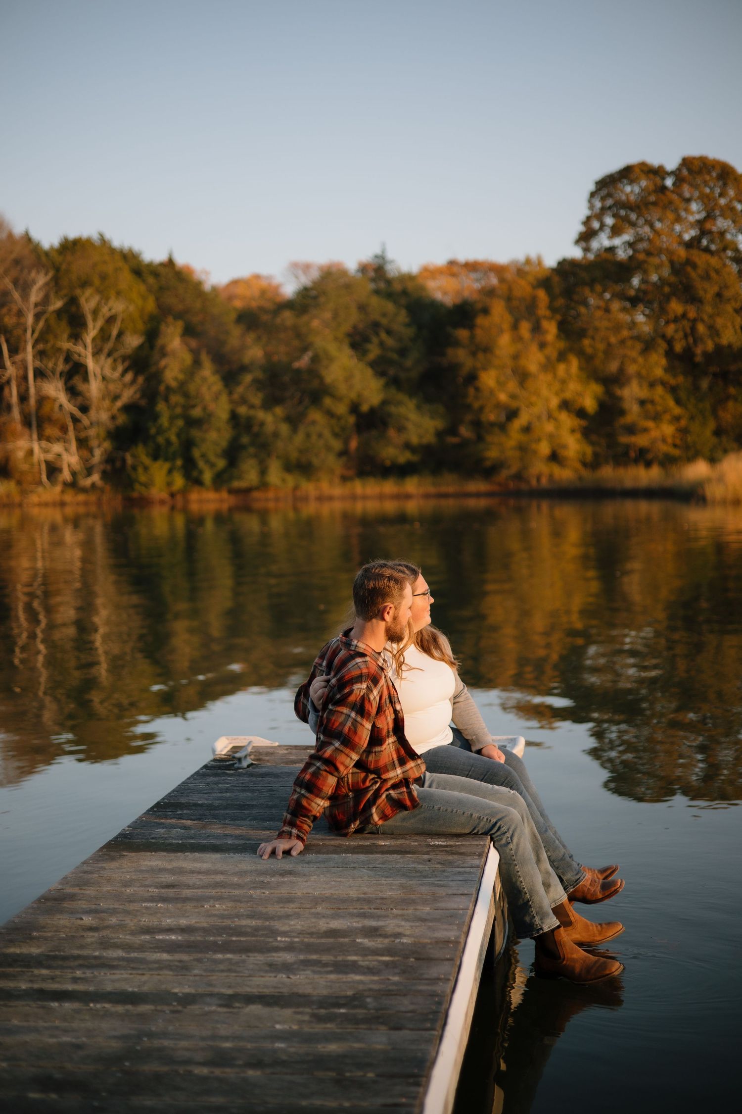 A couple sits together on a wooden dock overlooking a serene lake surrounded by autumn trees at sunset.