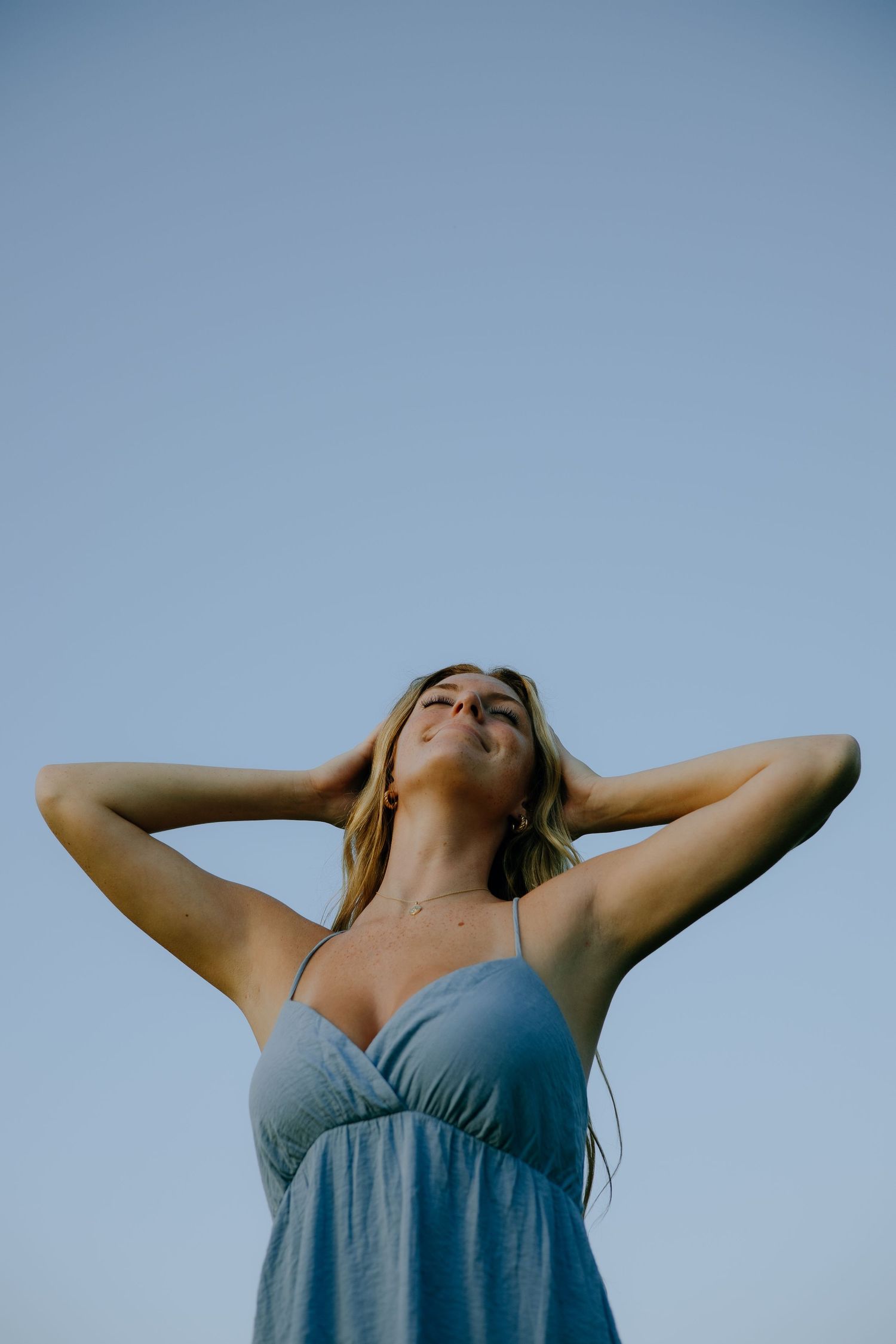 Person in blue tank top stretches arms up against bright sky background in relaxed pose.