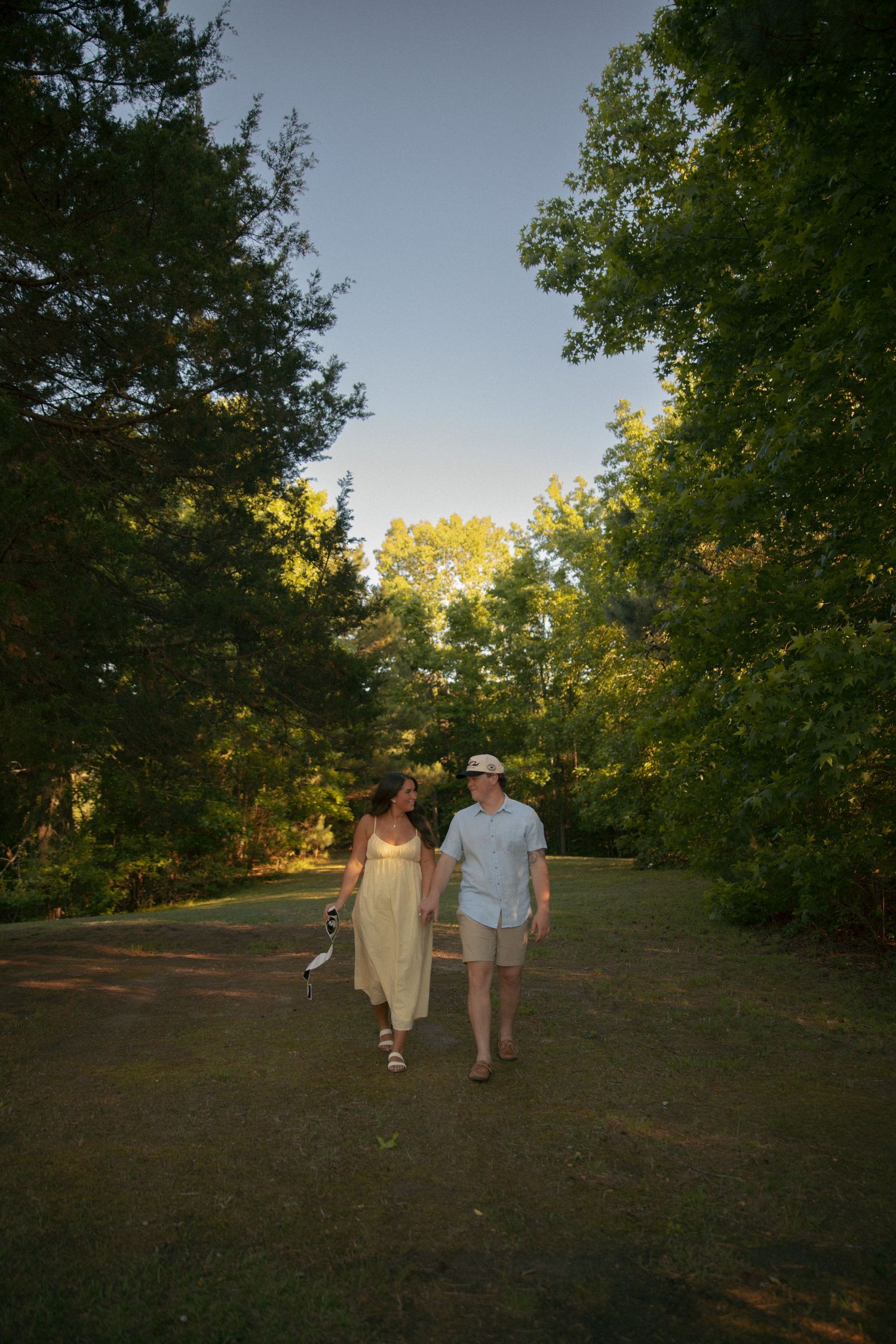 Couple holding hands walking on a sunlit garden path surrounded by lush green trees at golden hour.