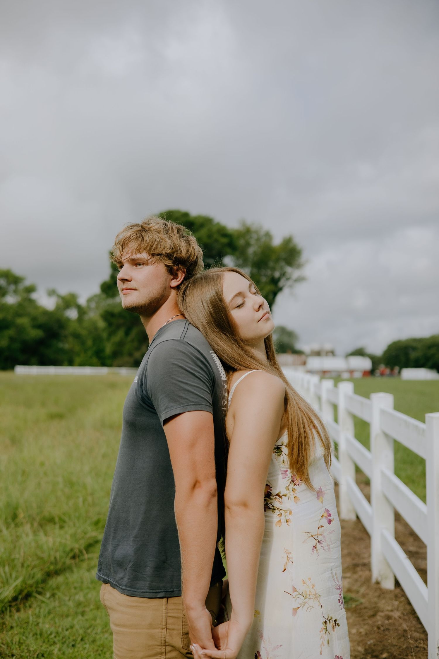 Young couple holding hands back-to-back by a white fence on a green farm field under cloudy skies.