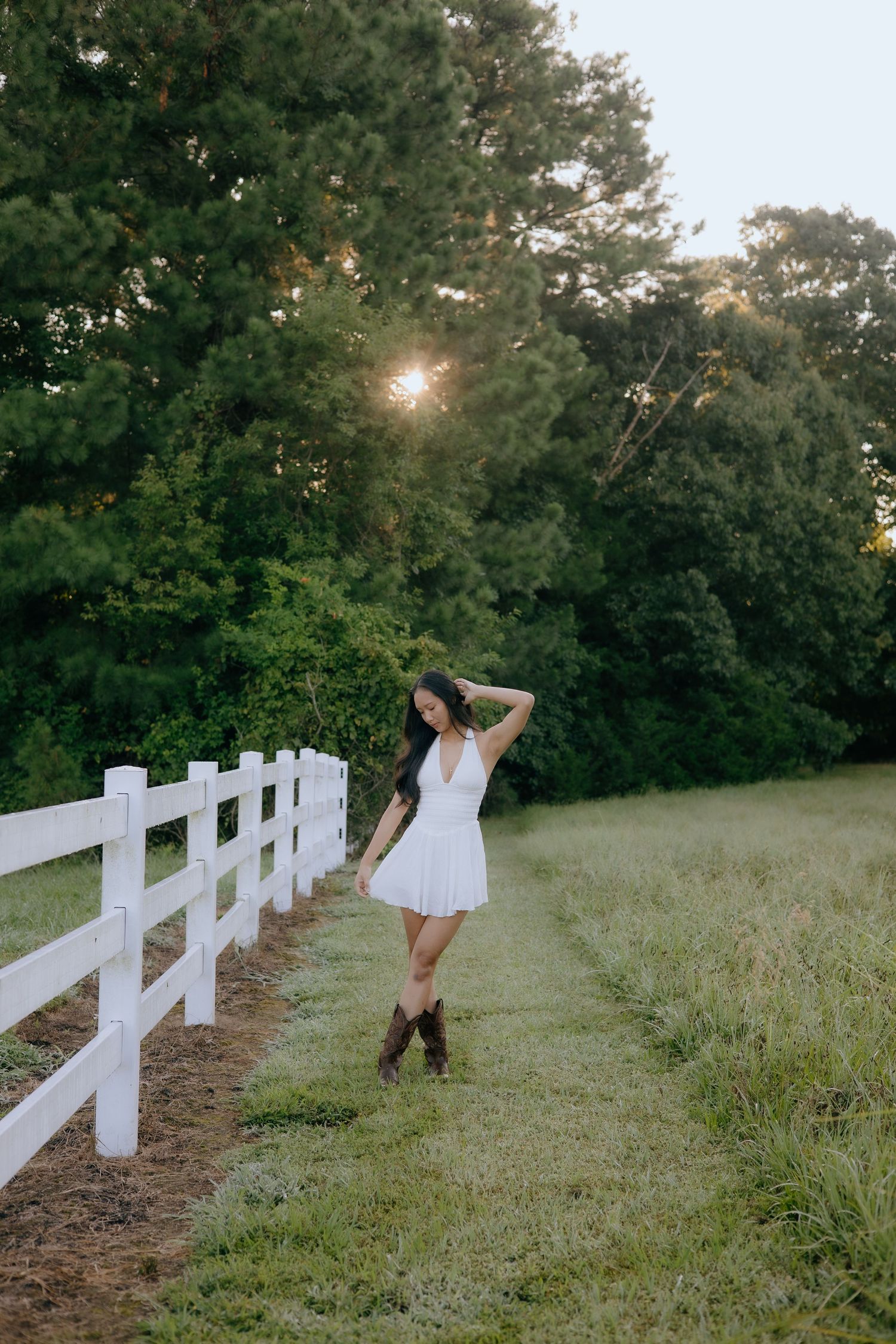 Young woman in white dress and cowboy boots poses by white fence at golden hour in lush green field.