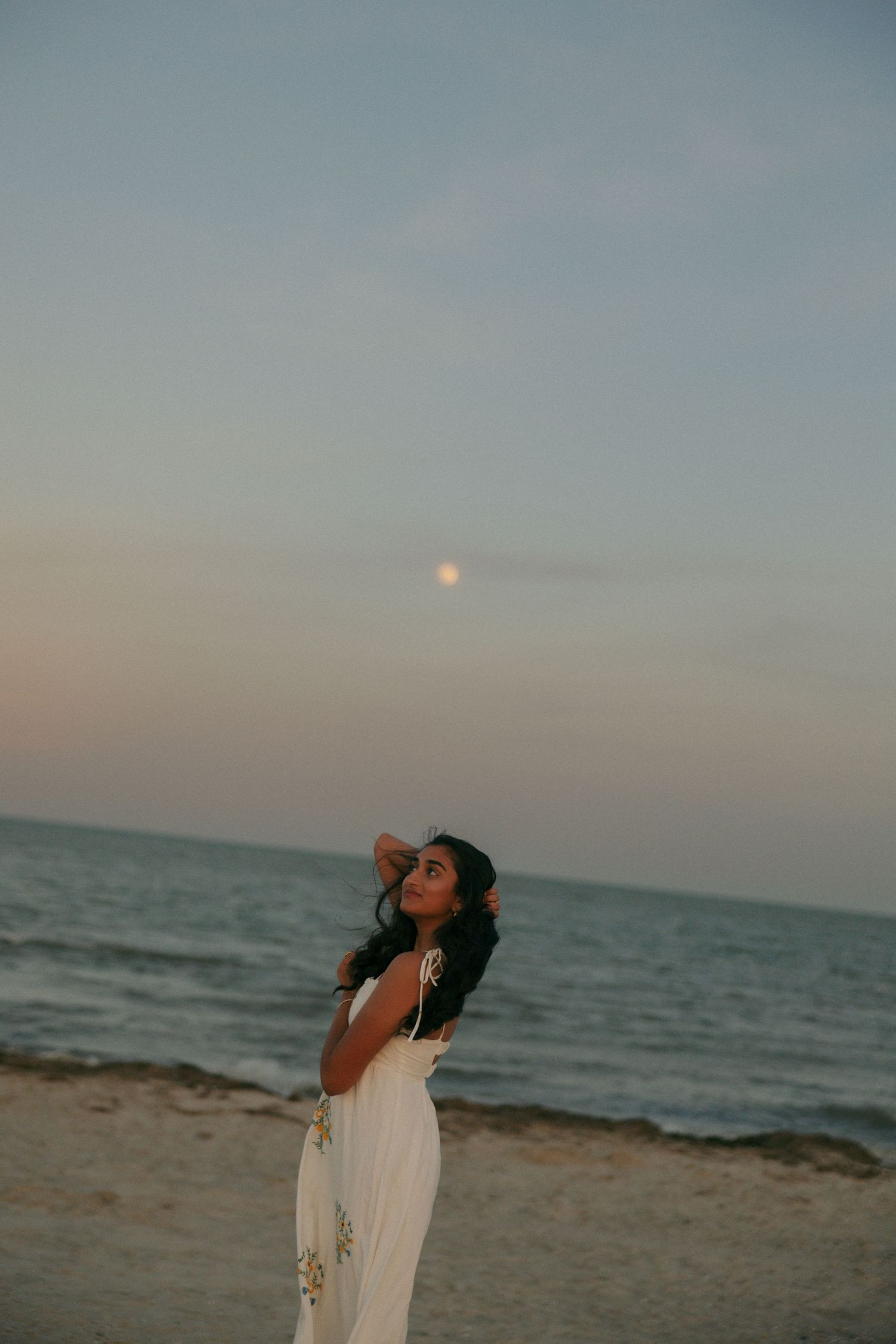 Woman in white dress gazing at the moon on a serene beach at dusk with calm ocean waves.