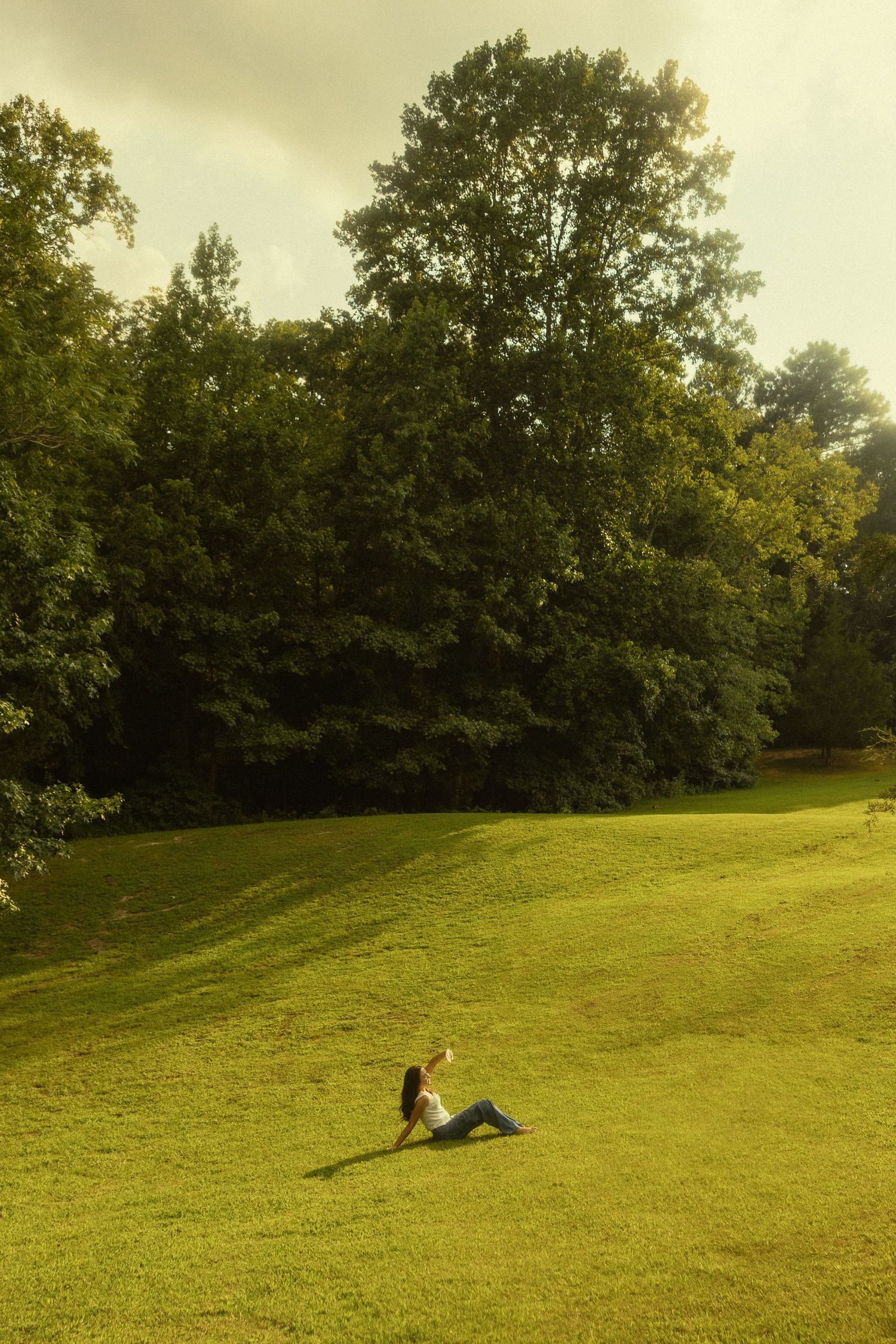 Woman sitting on lush green grass in a sunny park surrounded by tall trees on a bright day.