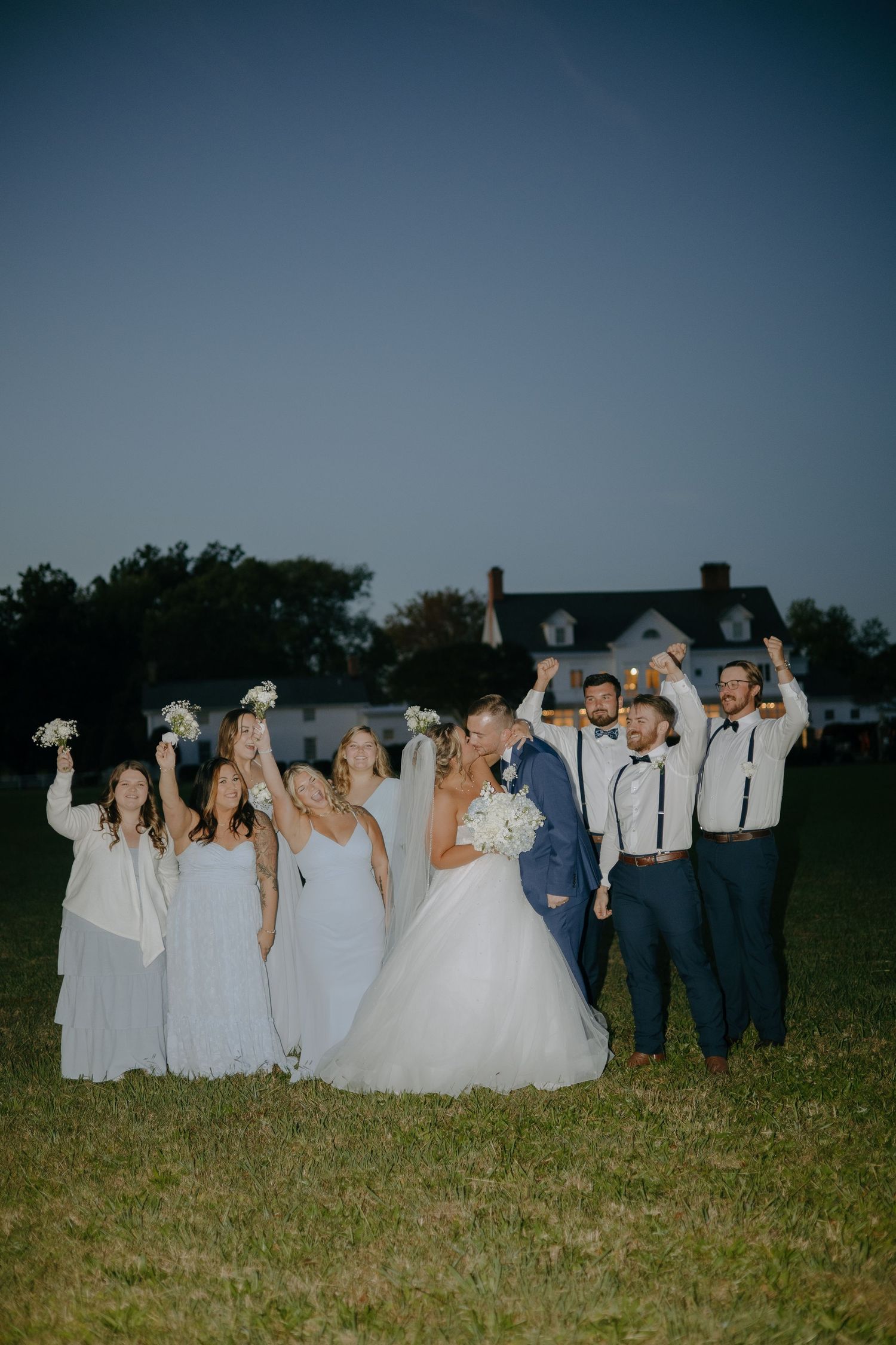 Wedding party poses together in a grassy field at dusk wearing navy blue and white attire.
