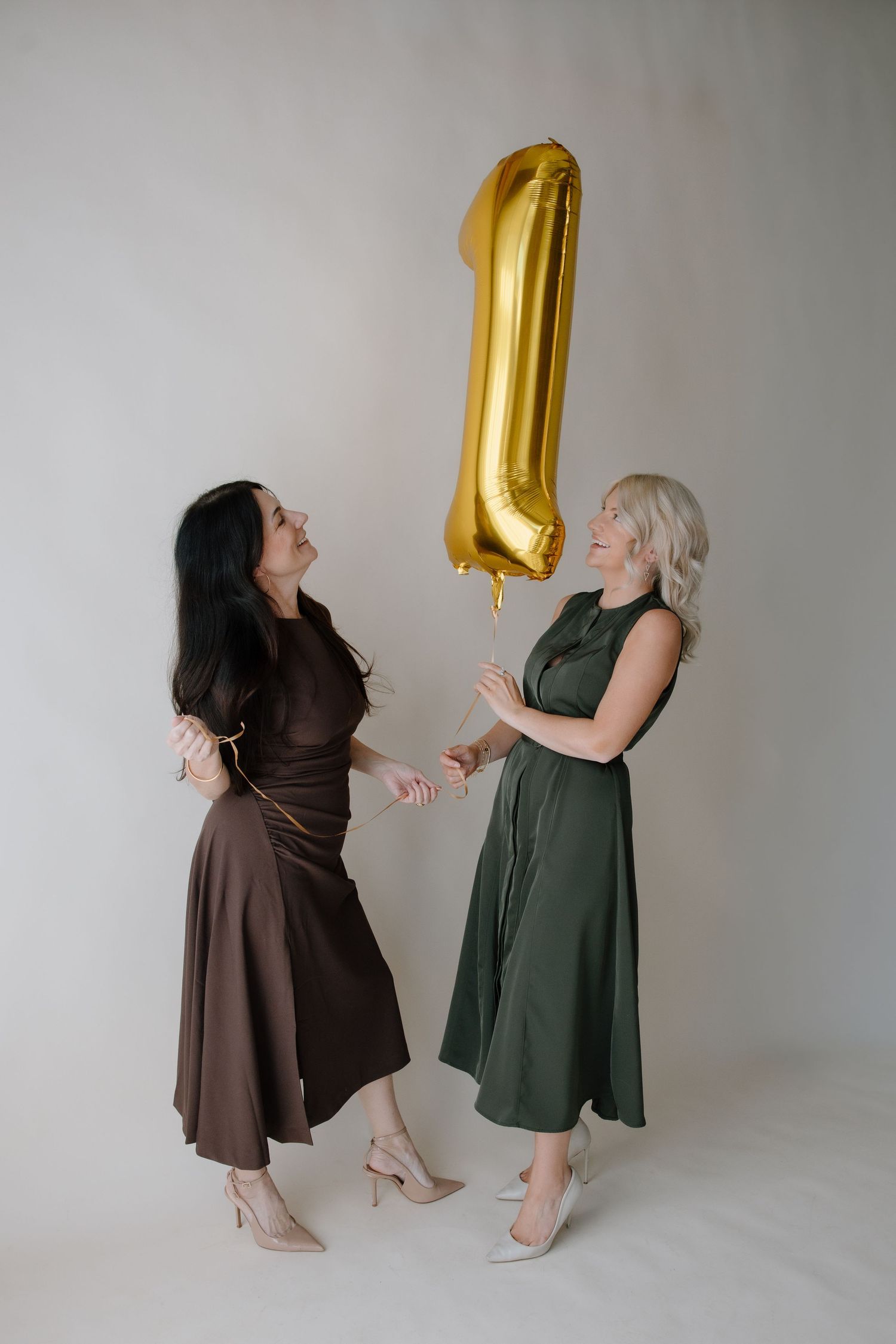Two people in contrasting outfits playfully interact with a gold number one balloon against a gray backdrop.