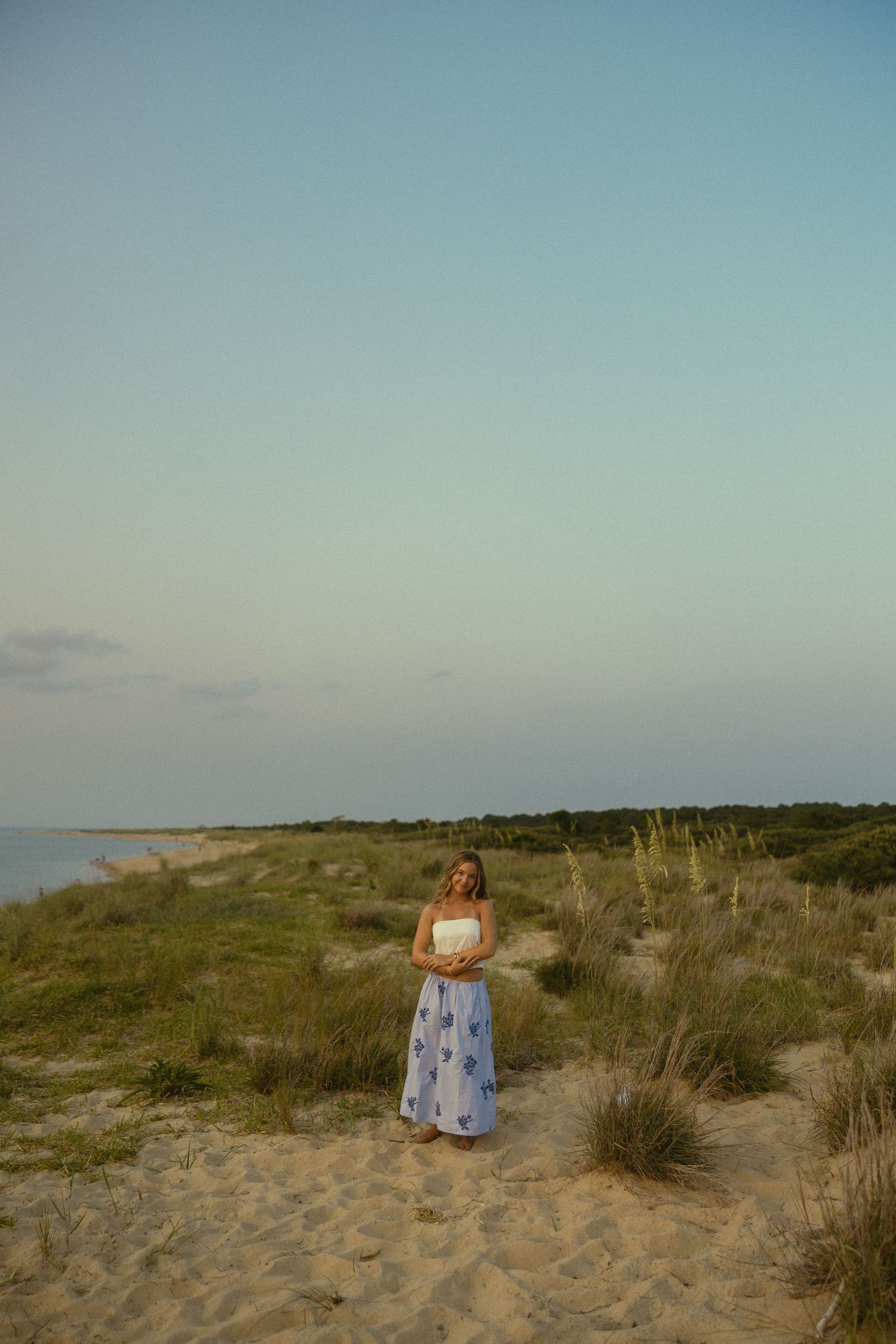 Woman in white top and blue floral skirt standing on sandy beach dunes at dusk with calm ocean horizon.