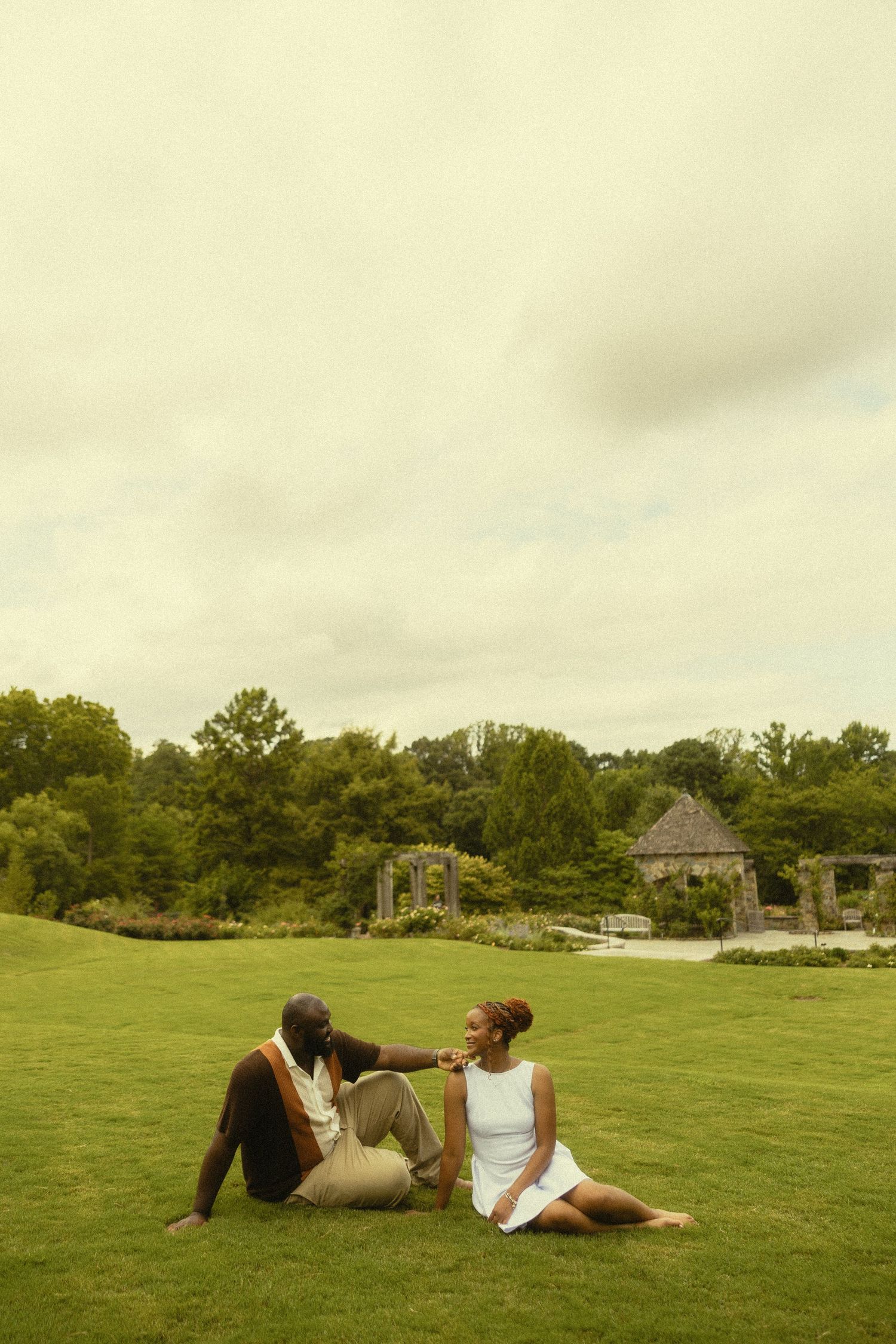 A couple sits on lush green grass in a garden park, sharing an intimate conversation near a gazebo.