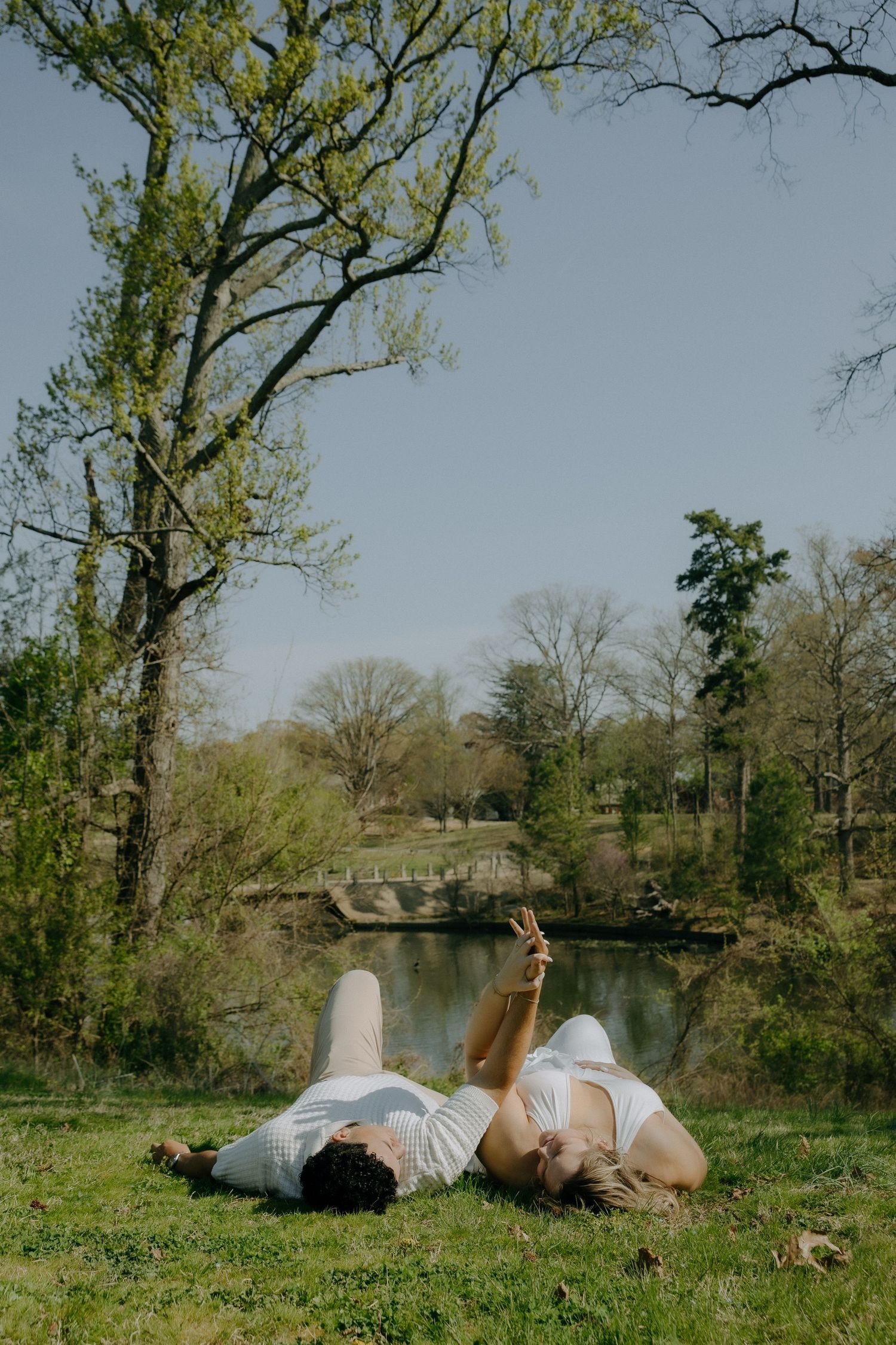 Couple lying on green grass by a peaceful pond, surrounded by spring trees in a sunny park.