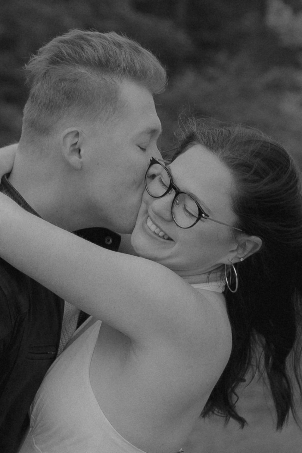 Black and white photo of a couple embracing, man kissing smiling woman with glasses on the cheek.