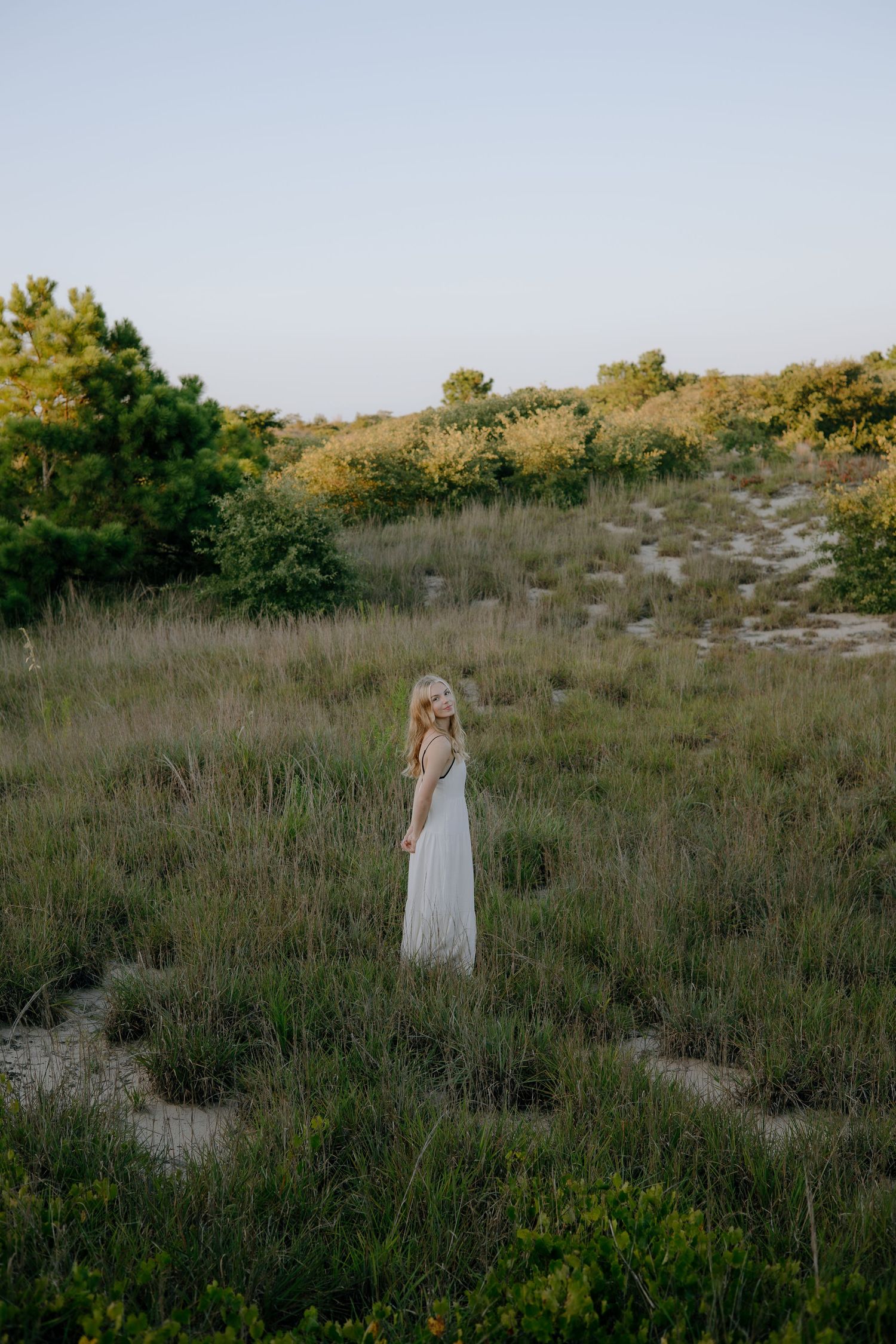 A young woman in a white dress stands in a lush green field surrounded by trees at dusk.