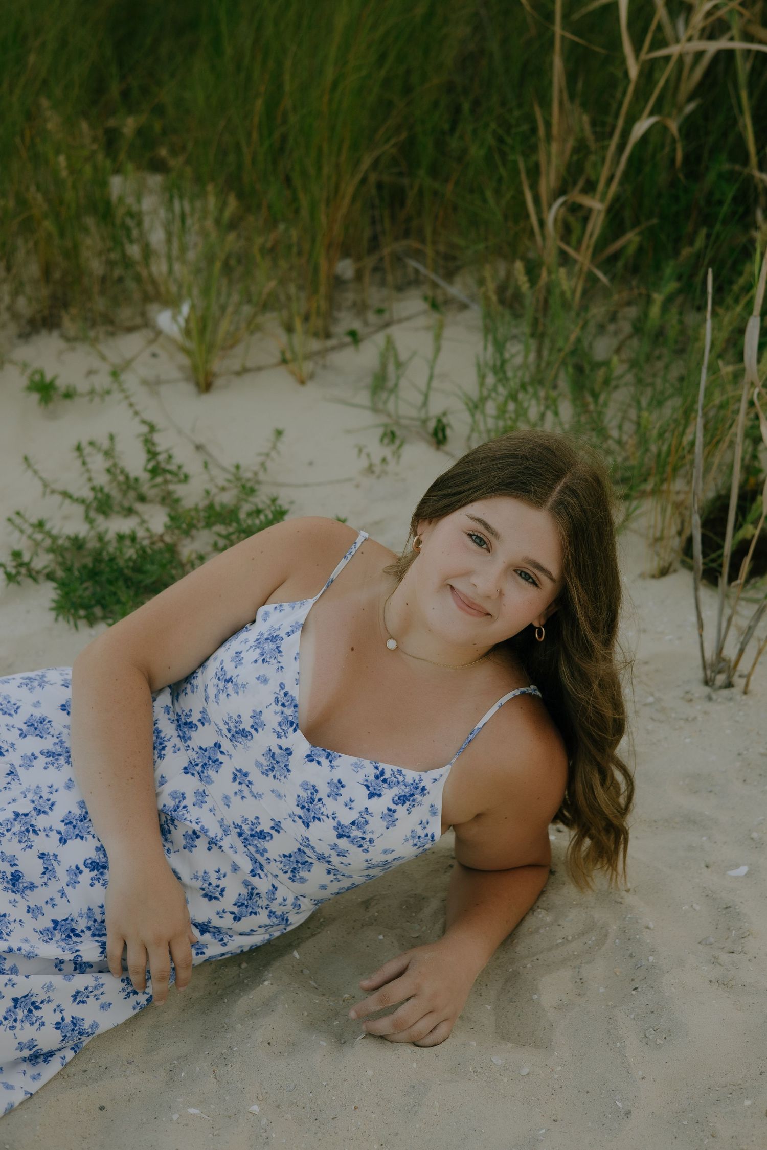 Person in a blue and white floral dress lying on sandy beach dunes with tall grass in the background.