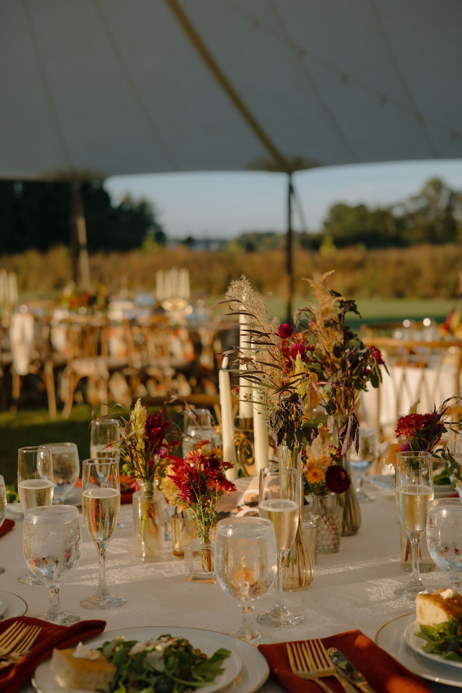 Elegant outdoor wedding reception table setting with candles and burgundy floral arrangements at golden hour.