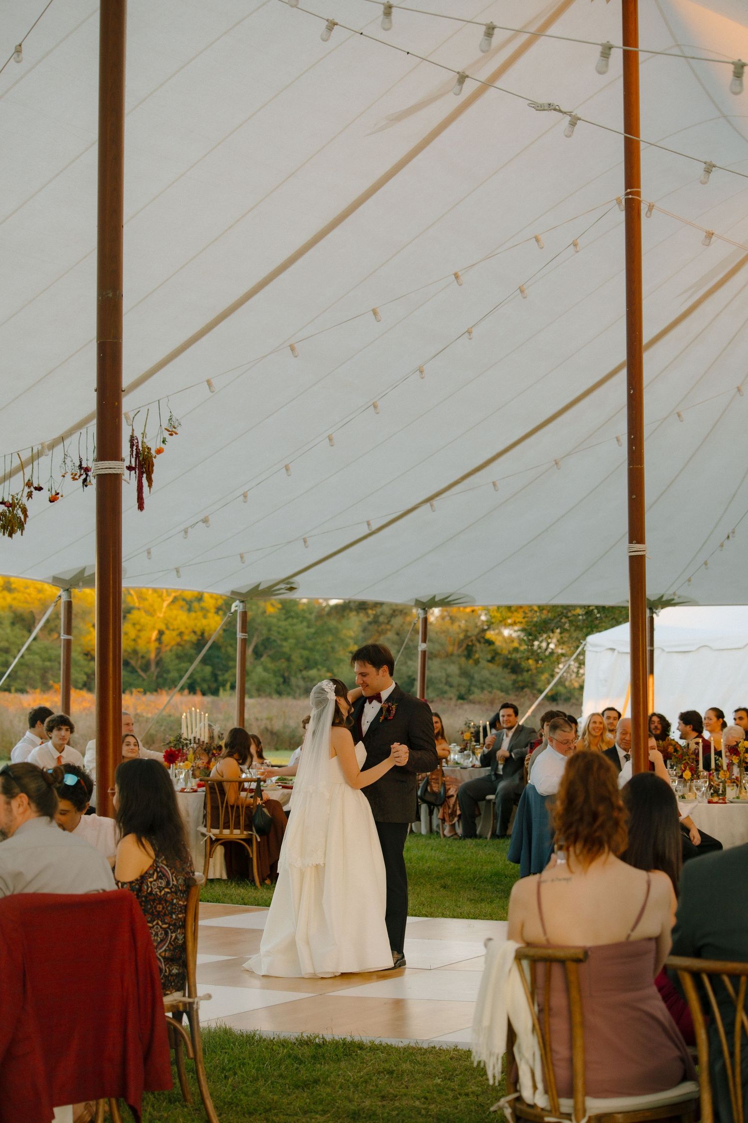 Wedding guests gather under an elegant white tent during an outdoor reception at sunset.