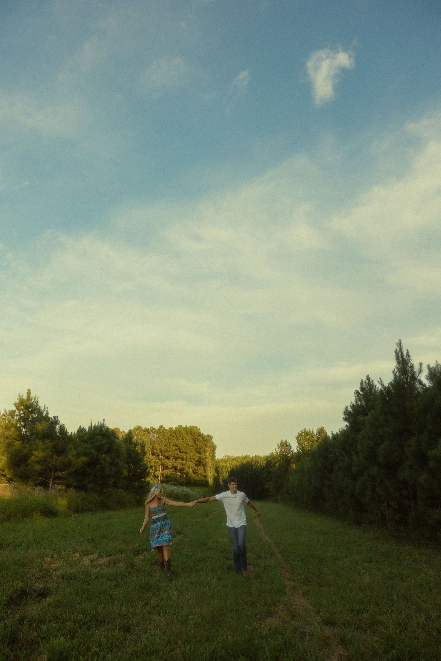Couple holding hands walking through a lush green field lined with trees under a blue sky at golden hour.