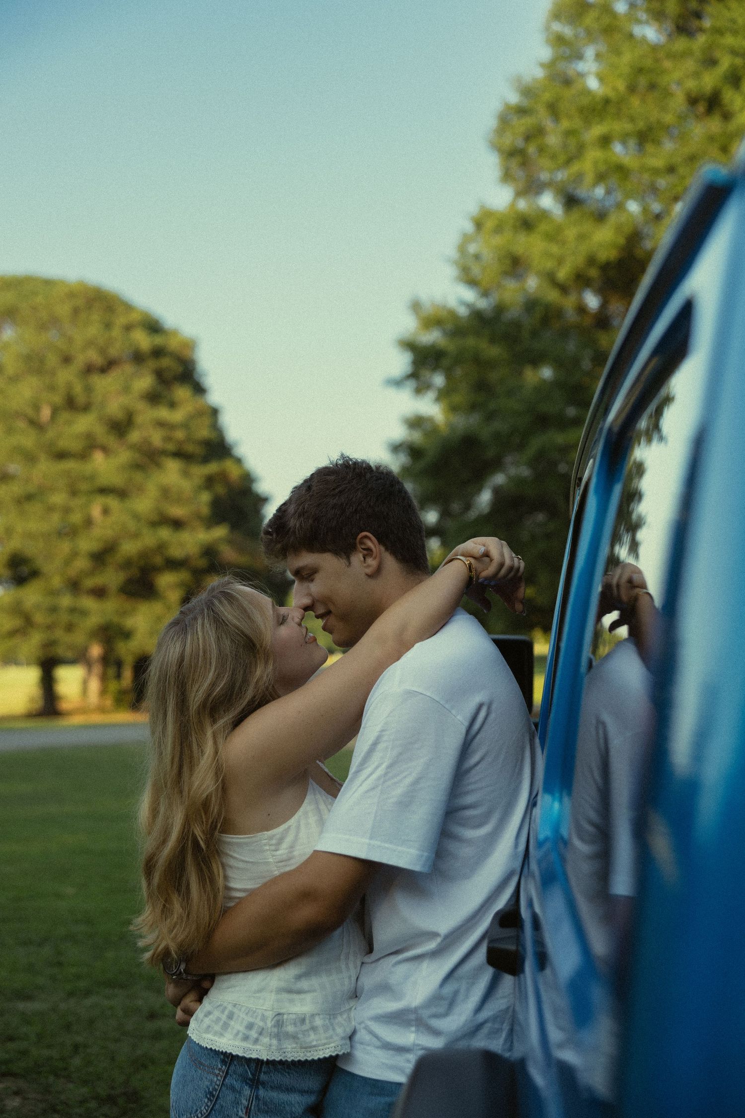 A young couple embracing and almost kissing beside a blue vehicle in a lush green park on a clear day.