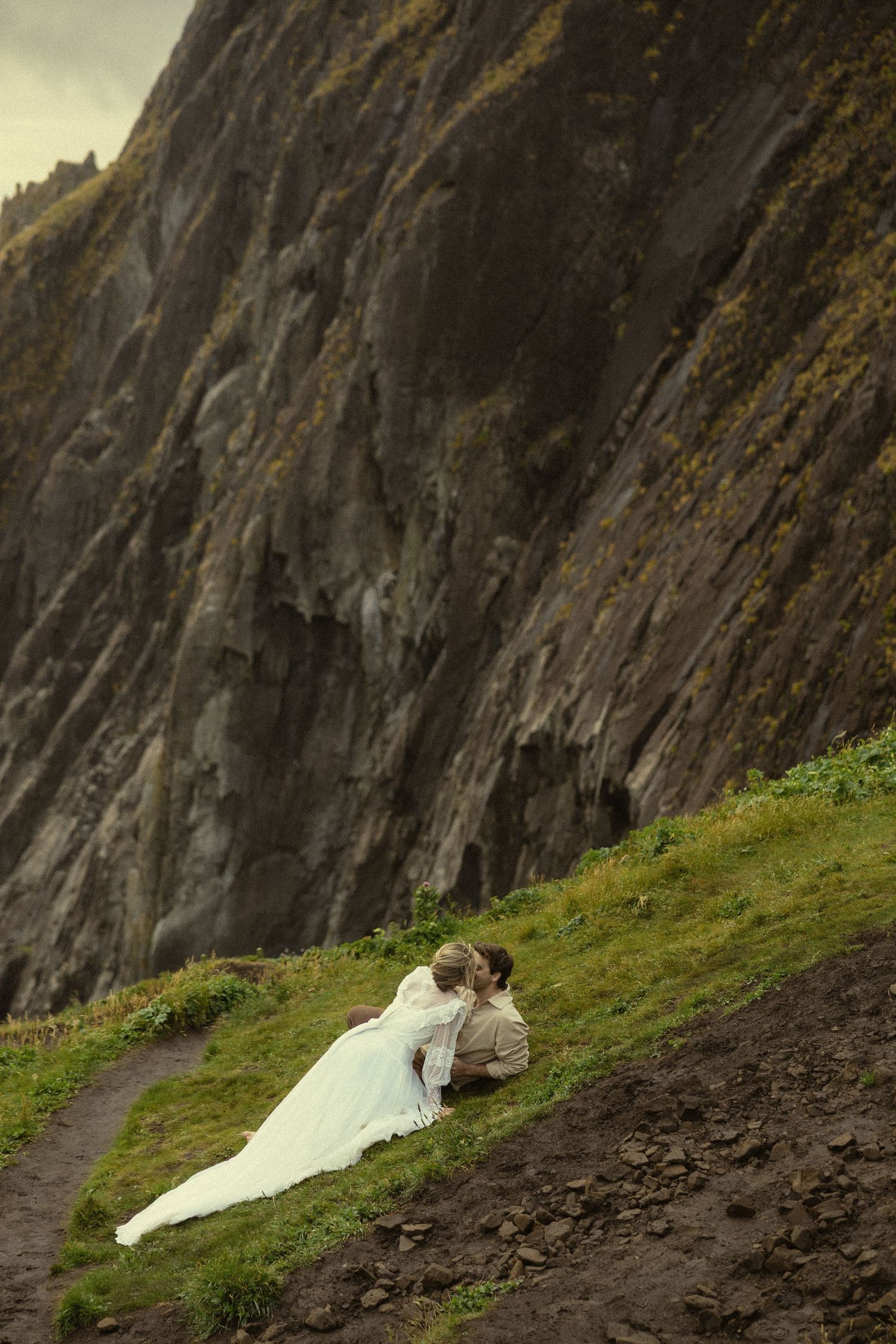 Bride and groom sitting together on a grassy hillside beside dramatic rocky cliffs on their wedding day.