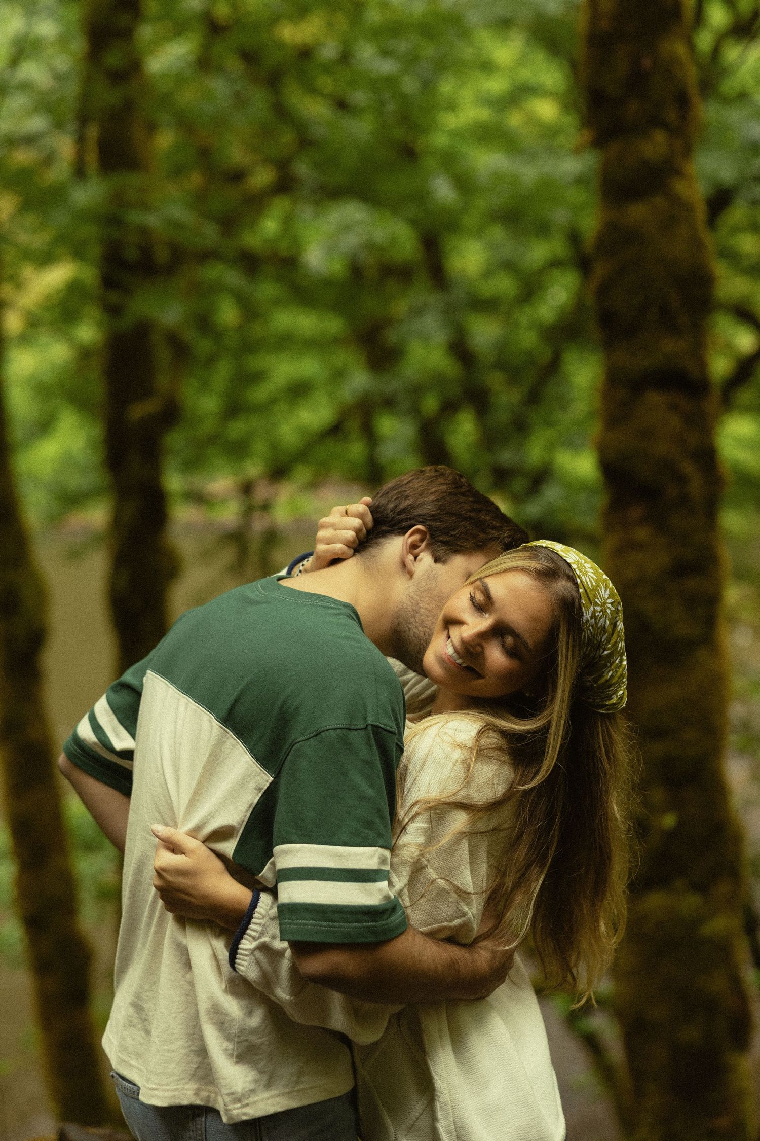Couple embracing romantically in a lush green forest, woman smiling with eyes closed, wearing a yellow headband.