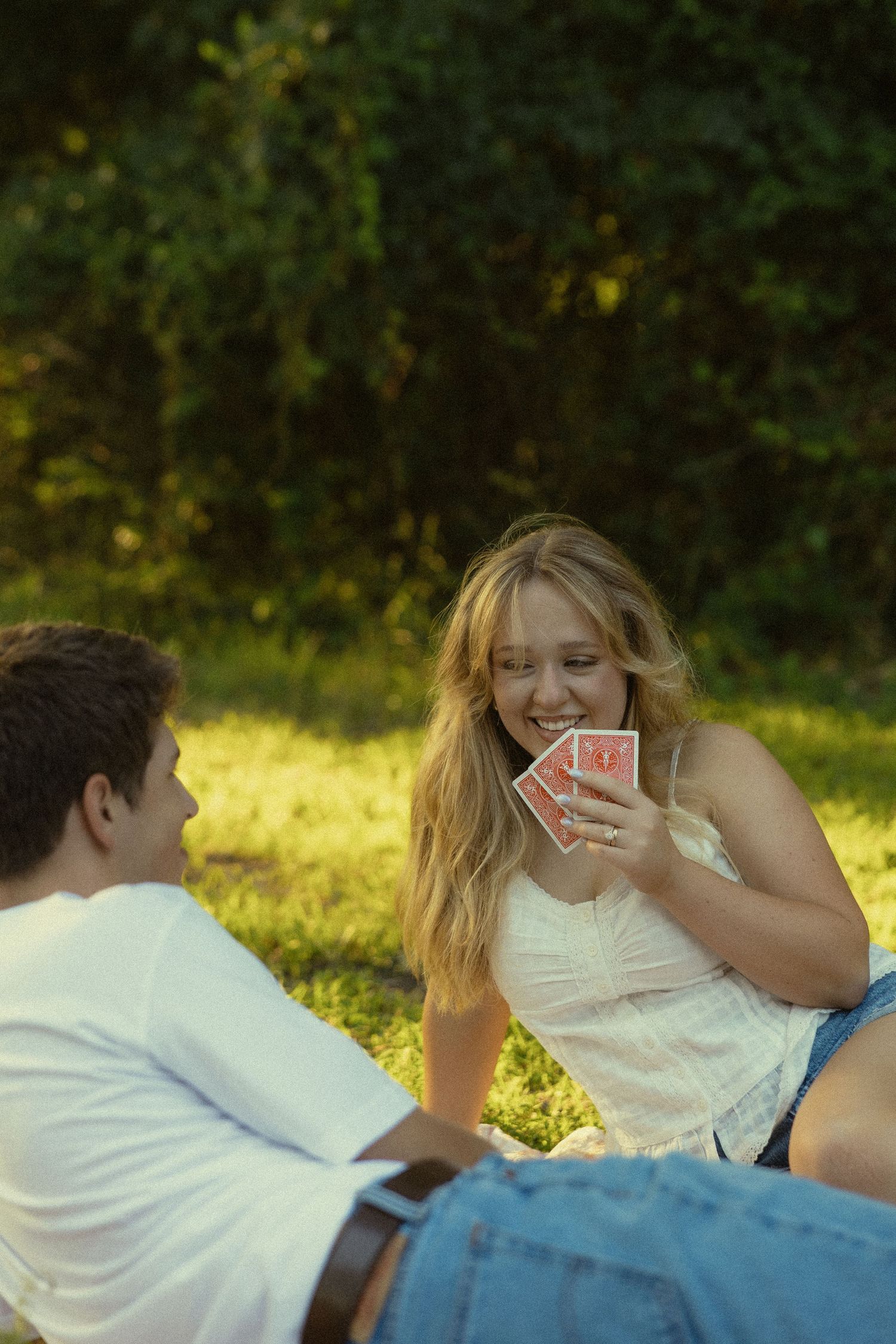 Young woman smiling and holding playing cards while sitting on grass with a young man during a sunny outdoor card game.