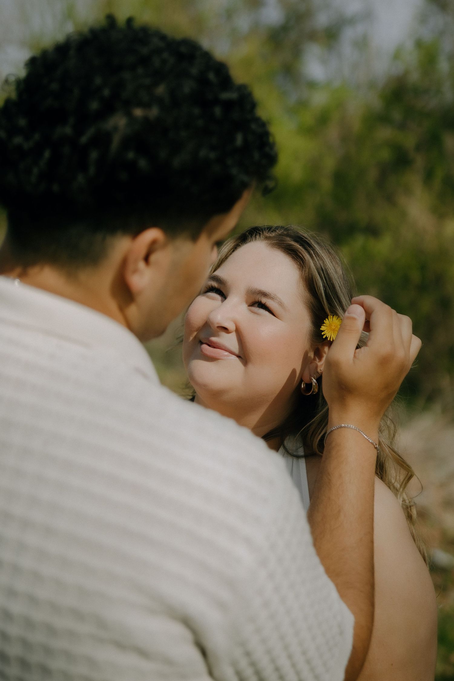 A man places a yellow flower behind a smiling woman's ear outdoors, in a tender romantic moment.