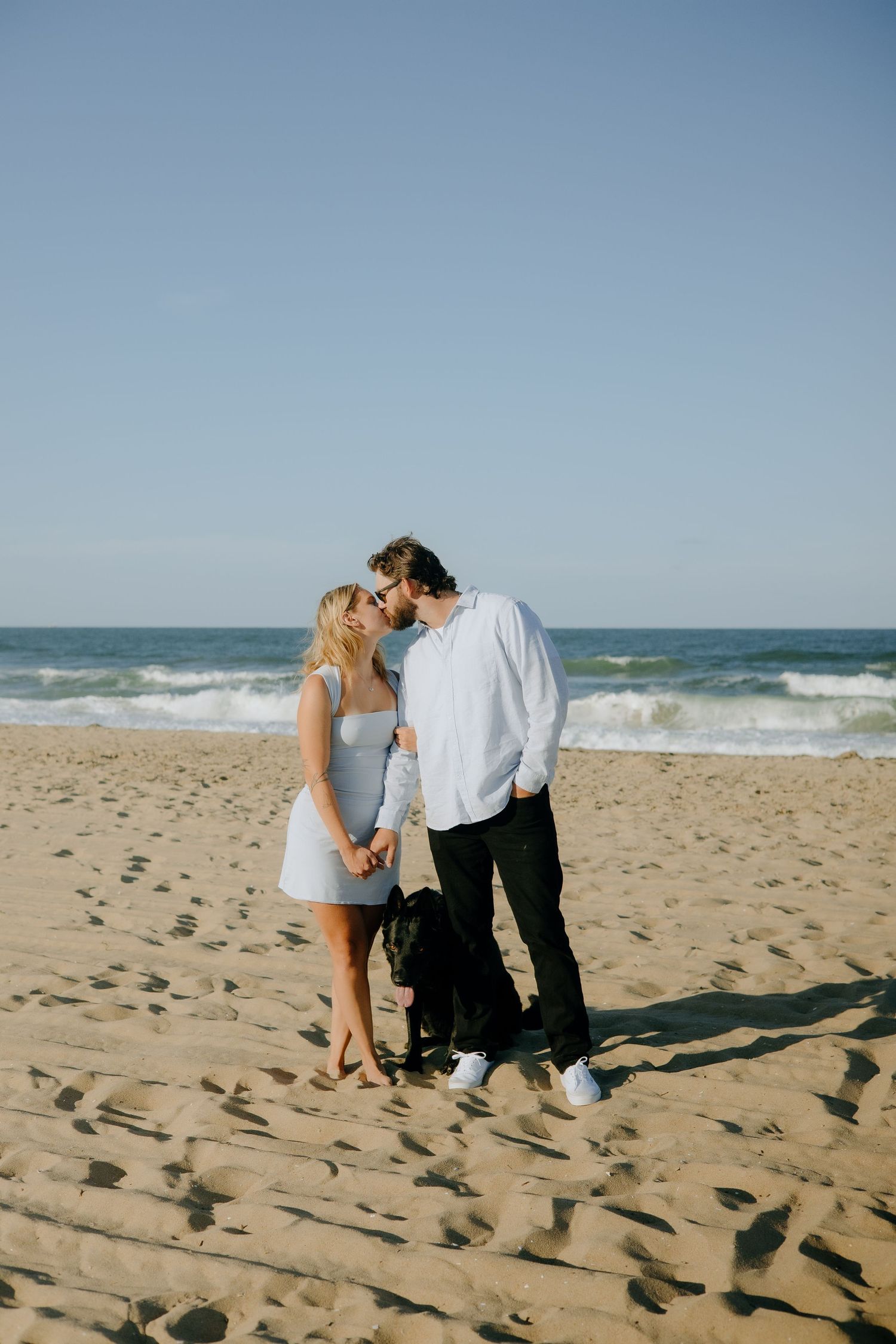Couple kissing on sandy beach with black dog, ocean waves in background, woman in white dress, man in white shirt.