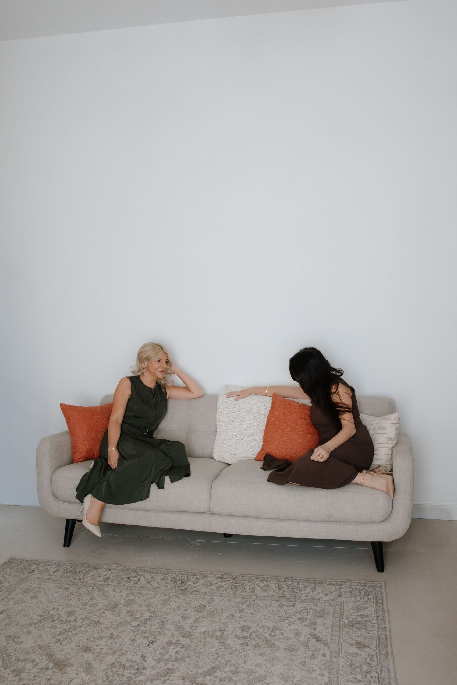 Two women sitting on a beige sofa with orange and white pillows, engaged in conversation in a minimalist white room.