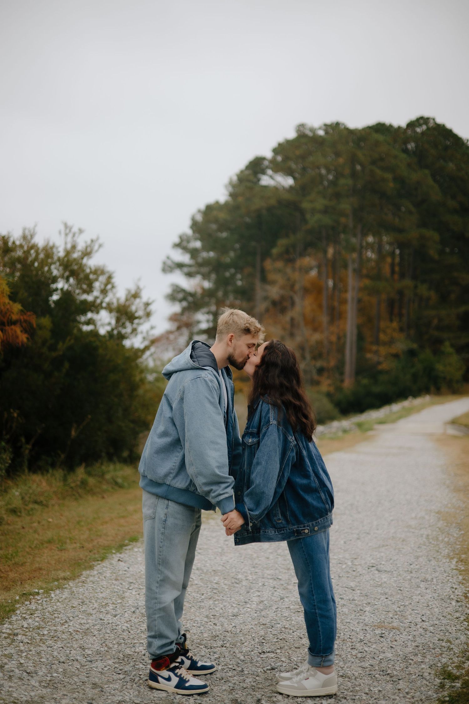 Young couple kissing on a gravel path surrounded by autumn trees, holding hands.