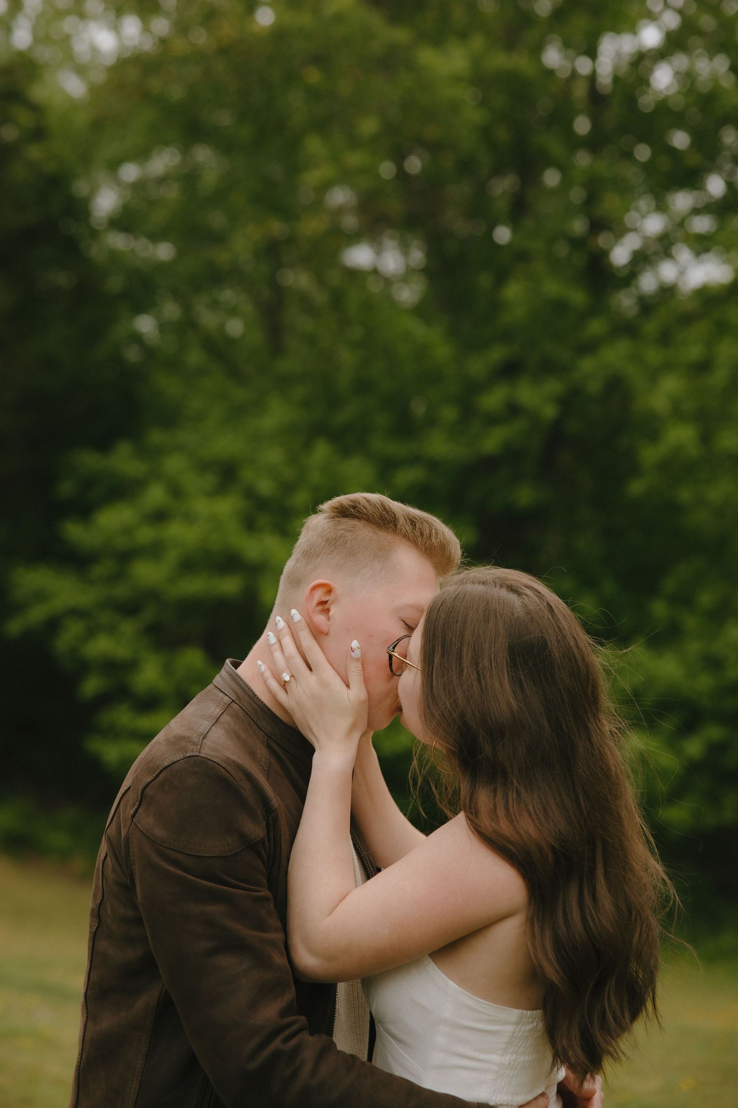 Couple sharing a romantic kiss outdoors, woman wearing engagement ring, surrounded by lush green trees.