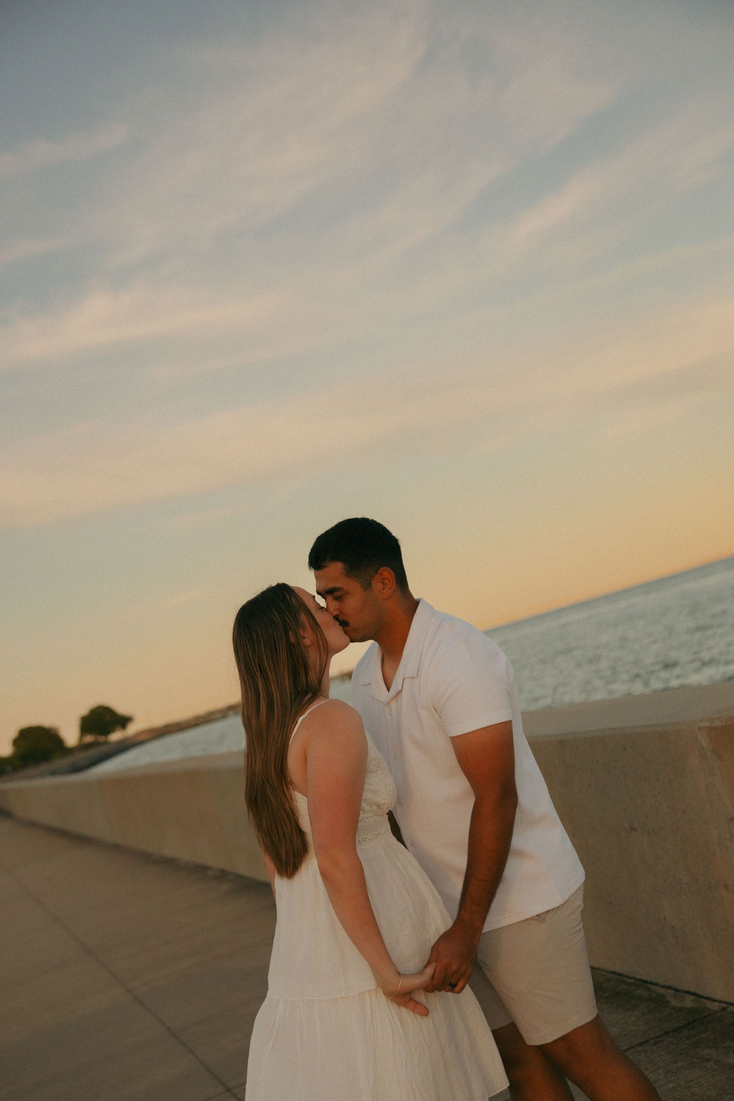 Couple sharing a romantic kiss at sunset by the waterfront, woman in white dress, man in white shirt and khaki shorts.