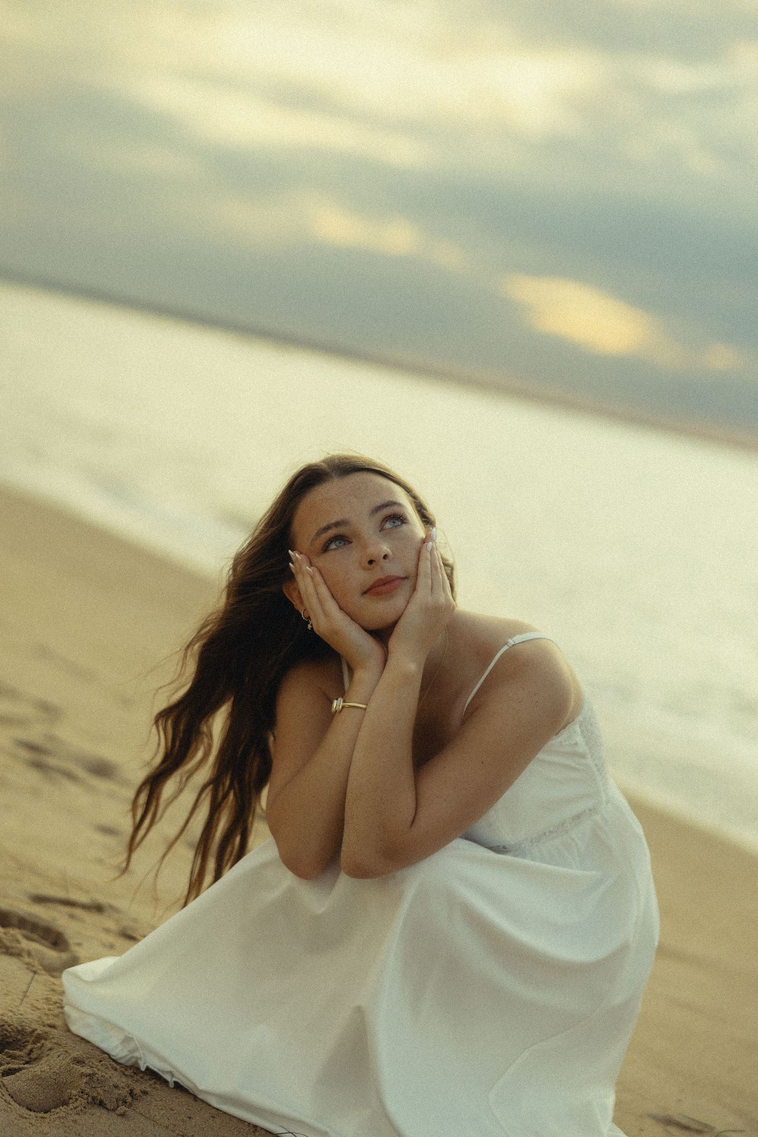 A woman in a white dress sits on a sandy beach at sunset, gazing upward with hands framing her face.